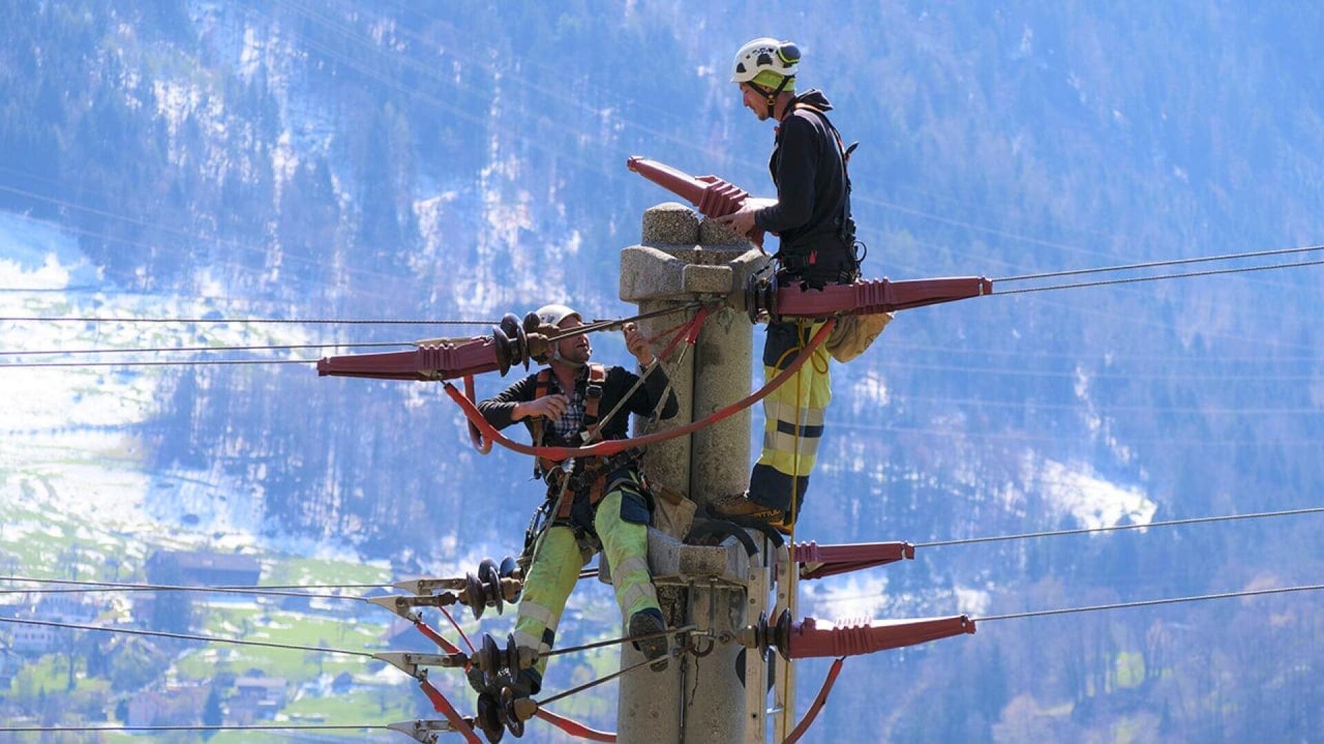 Zwei Arbeiter eines Energieversorgungsunternehmens in Schutzkleidung reparieren oder überprüfen Stromleitungen auf einem hohen Strommast, mit einer bergigen Landschaft im Hintergrund.