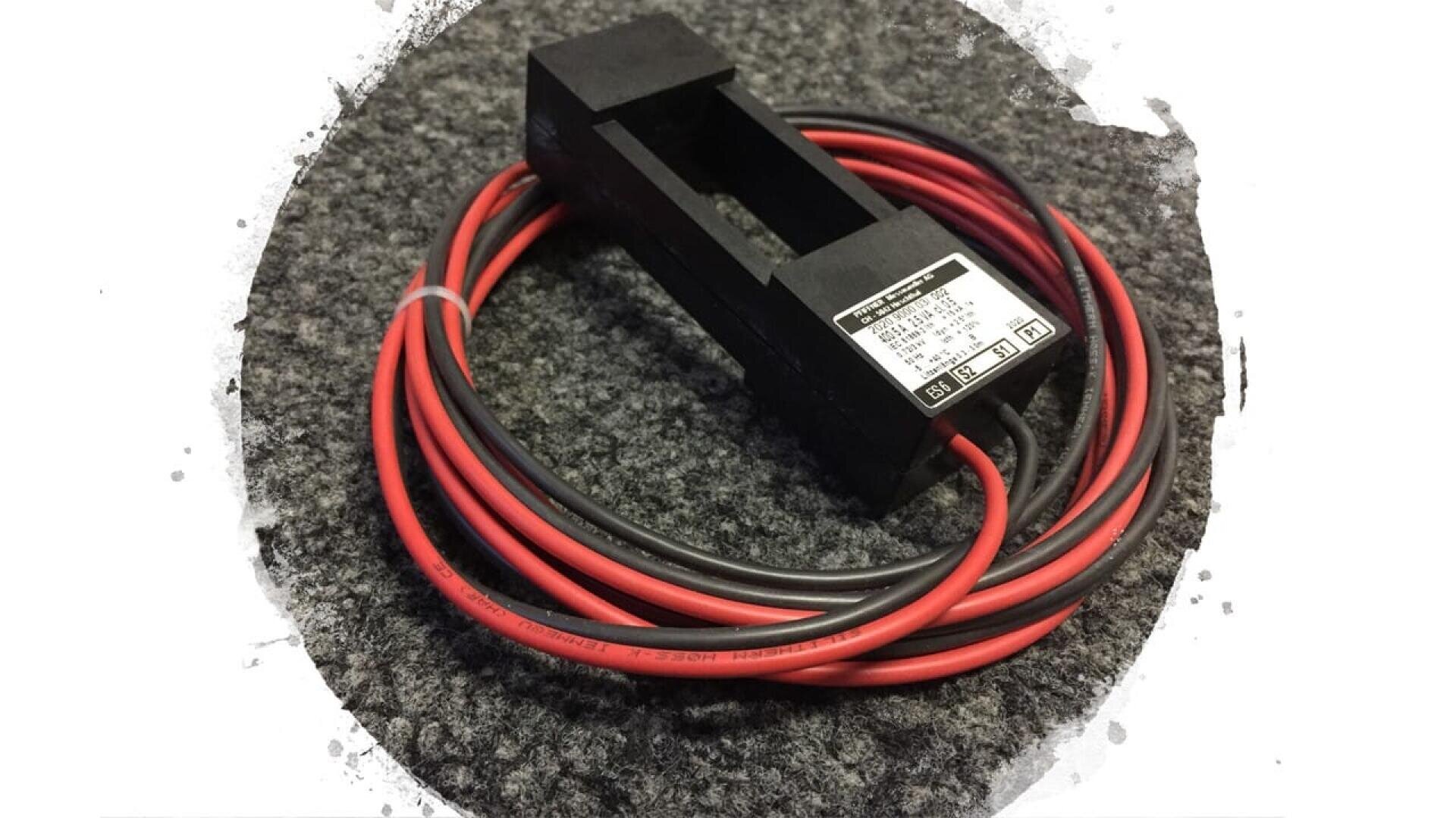 Ein schwarzer elektronischer Stromsensor mit einer rechteckigen Öffnung, der an roten und schwarzen Drähten befestigt ist, die ordentlich gewickelt auf einer strukturierten grauen Oberfläche liegen.