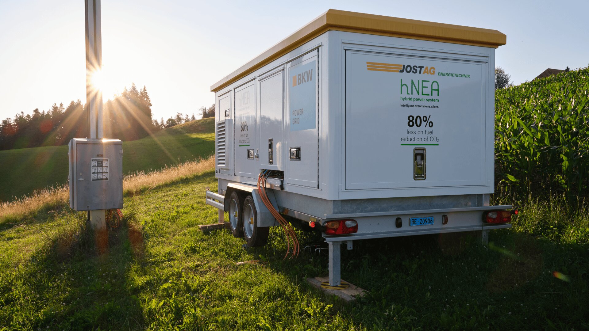 Eine weiße mobile Wasserstofftankanlage mit der Aufschrift Jostag hNEA steht bei Sonnenuntergang auf einer Wiese in der Nähe eines Maisfeldes. Sie ist über Kabel mit einem nahe gelegenen Stromkasten verbunden; im Hintergrund sind Bäume und Sonnenlicht zu sehen.