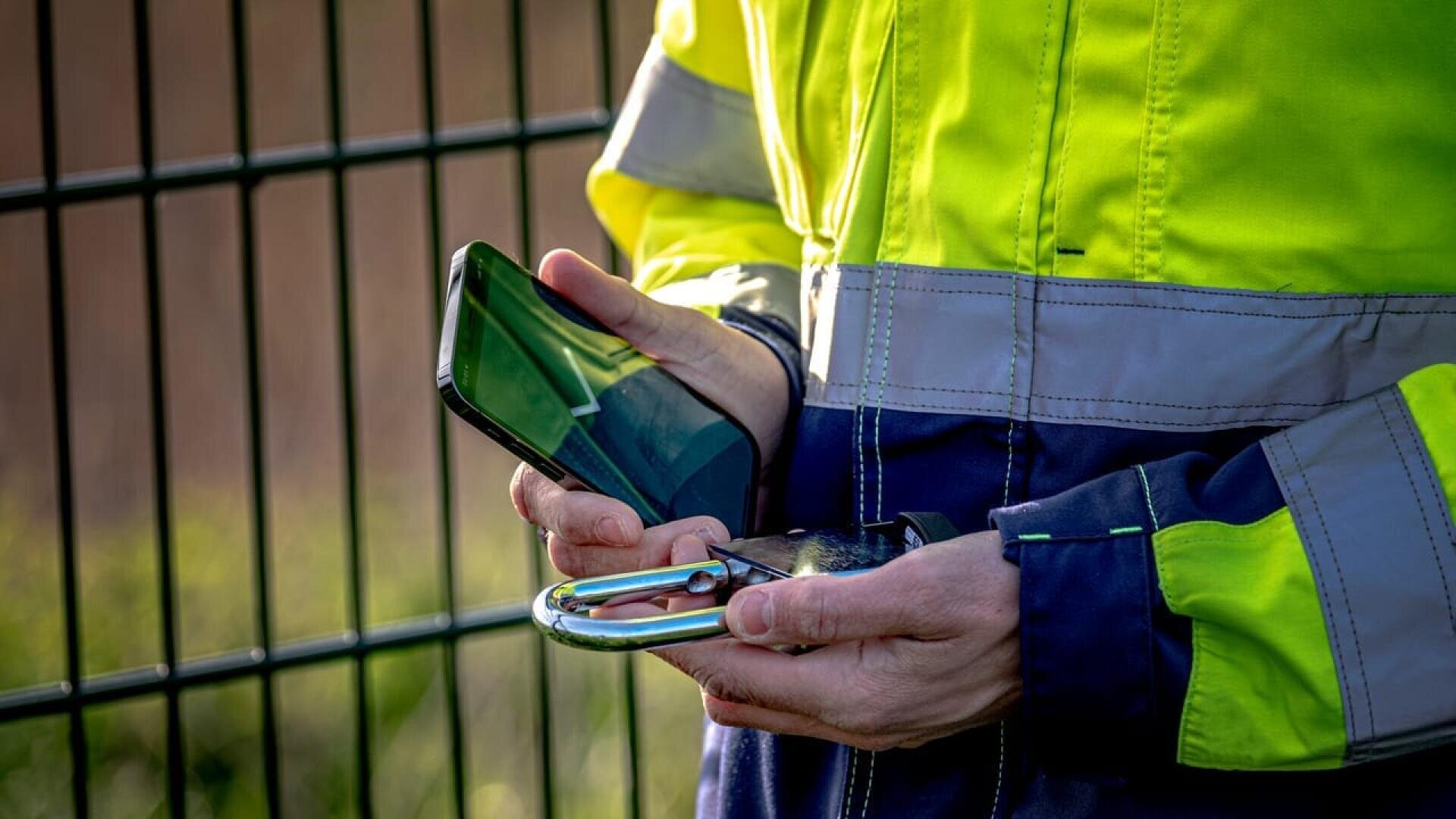 Eine Person in einer Warnweste hält ein Smartphone und ein großes Vorhängeschloss in der Hand, während sie vor einem Metallzaun steht.