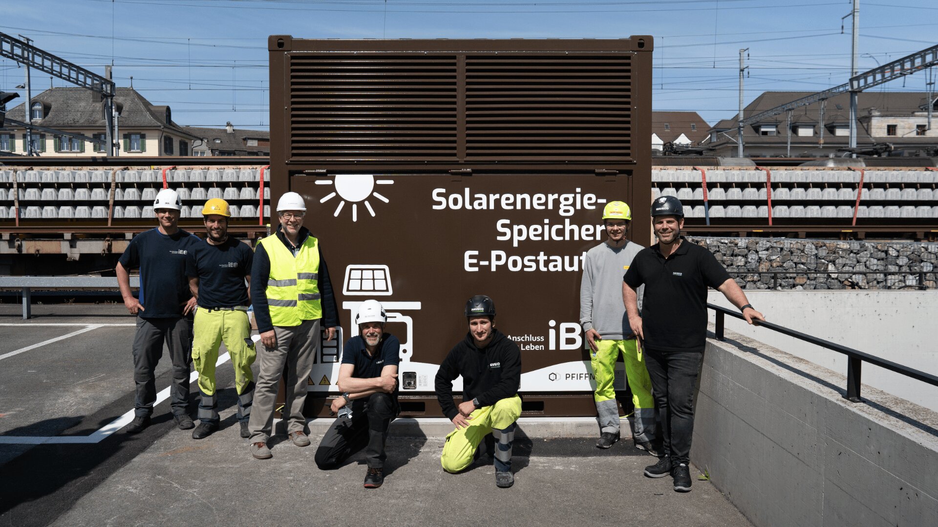 Sieben Bauarbeiter in Schutzkleidung posieren vor einem großen braunen Container mit der Aufschrift Solarenergie-Speicher, E-Postauto. Im Hintergrund sind Gleise und Gebäude zu sehen.