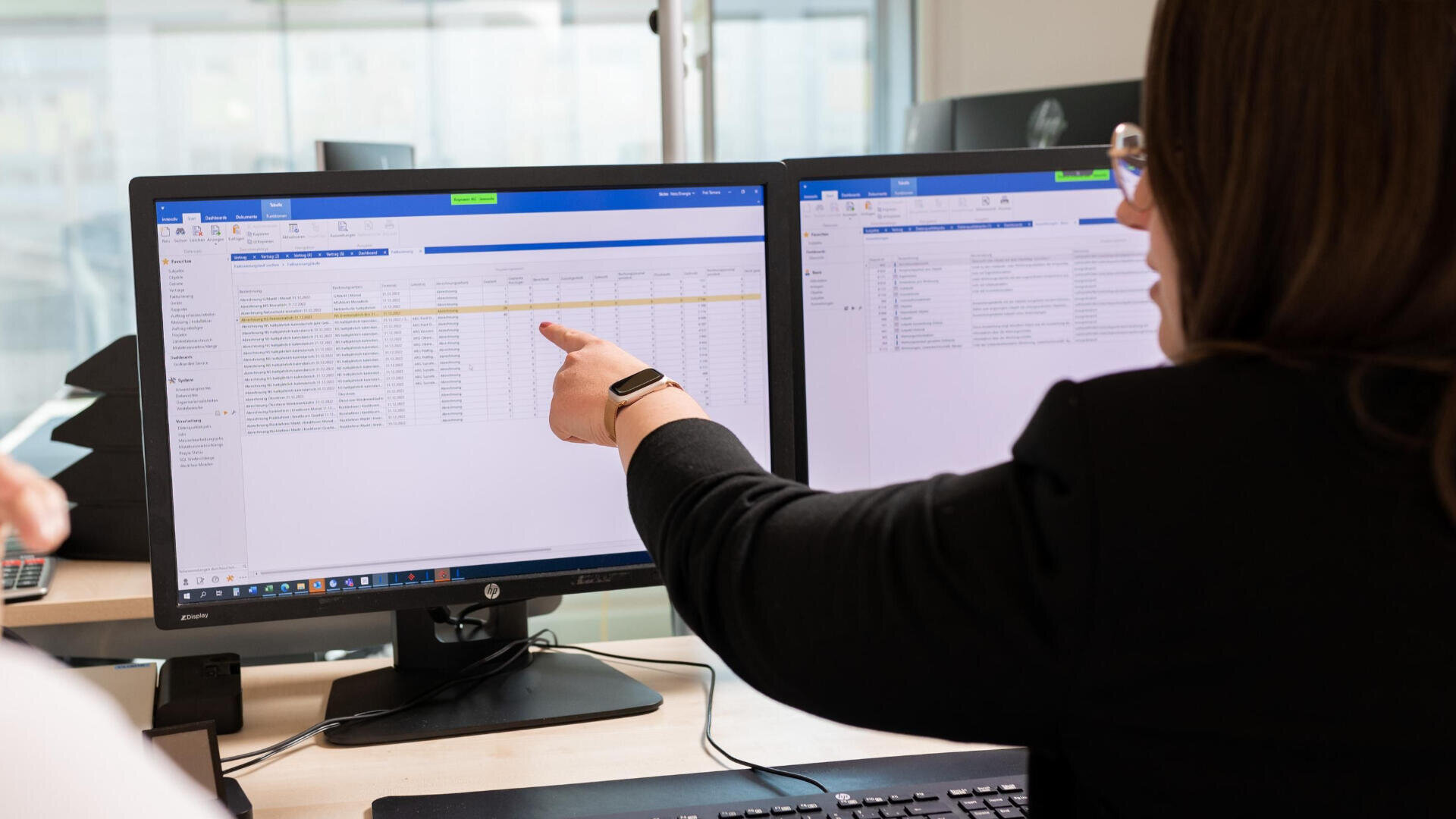 Eine Person, die eine Brille trägt, zeigt auf Daten auf einem Computermonitor, während sie an einem Schreibtisch mit zwei Bildschirmen sitzt, auf denen Tabellenkalkulationen oder Datenbankinformationen in einer Büroumgebung angezeigt werden.
