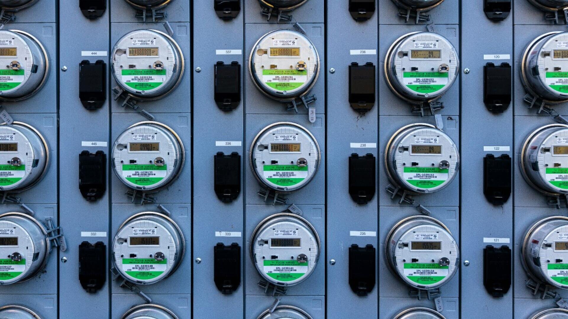 A close-up view of multiple electric utility meters arranged in a grid on a gray panel, each meter displaying digital readings and featuring green labels at the bottom.