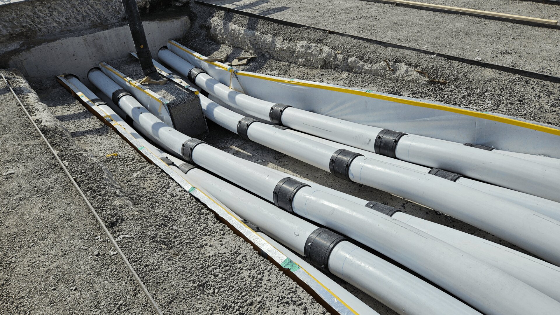 Große graue Versorgungsrohre sind auf einer Baustelle fein säuberlich in Gräben angeordnet, mit schwarzen Befestigungselementen gesichert und von Kies und unfertigem Beton umgeben.
