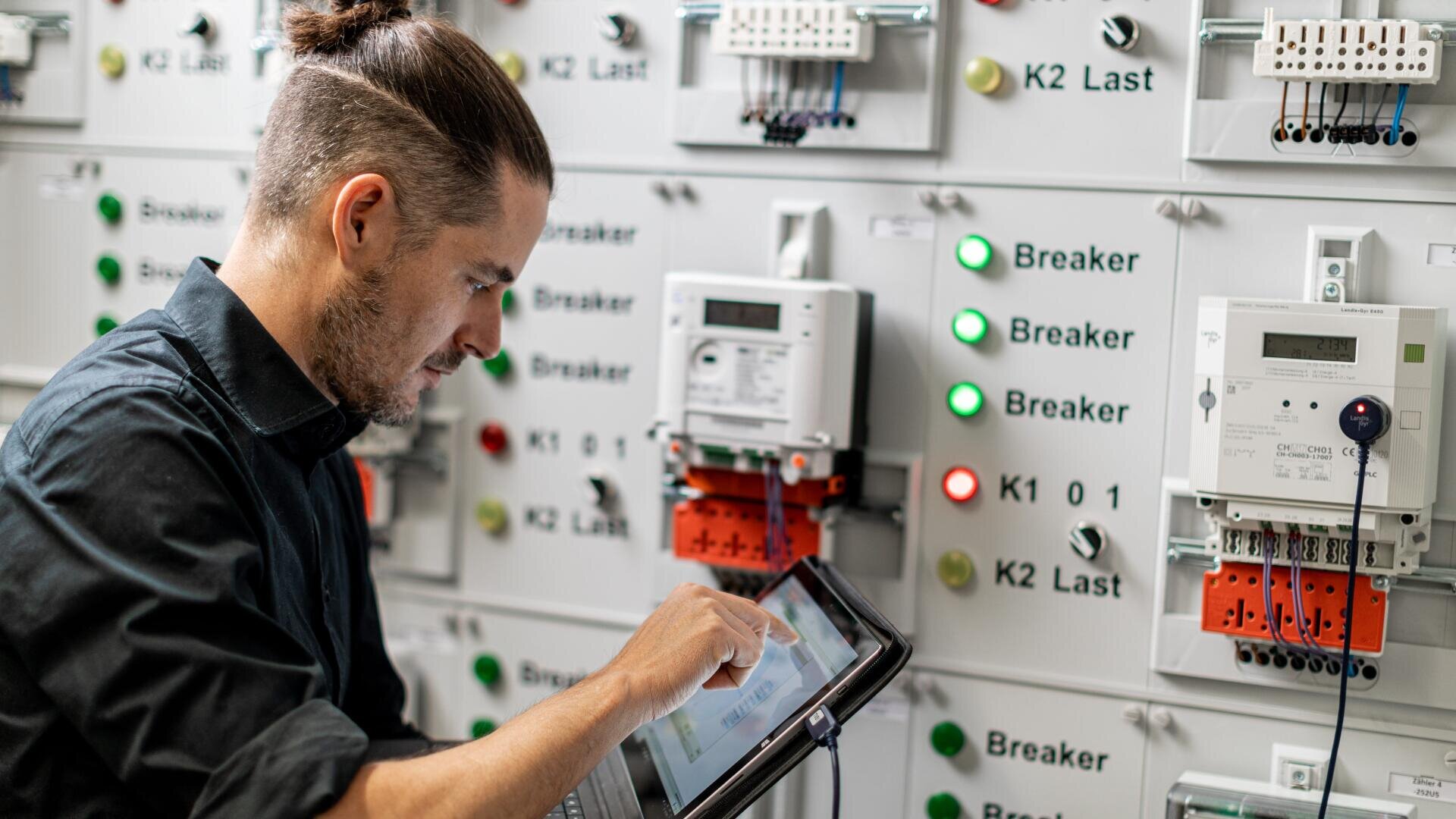 Ein Mann, der ein Tablet benutzt, prüft eine elektrische Schalttafel mit beschrifteten Unterbrechern, Messgeräten, Tasten und Anzeigeleuchten in einer technischen oder industriellen Umgebung.