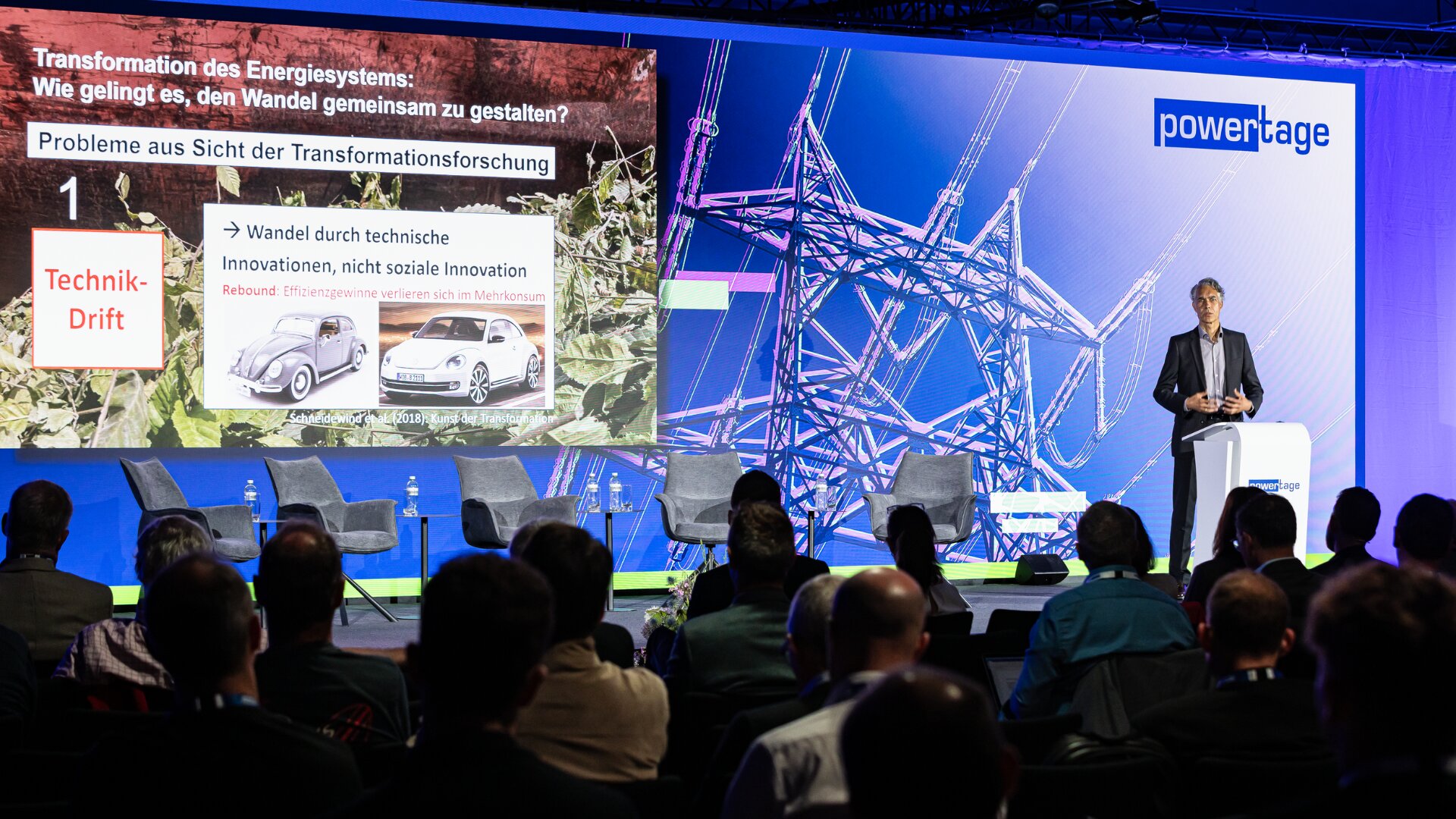 Ein Redner hält auf der Bühne einer Konferenz einen Vortrag, auf dem eine Folie über die Energiewende mit Bildern von alten und modernen Autos gezeigt wird. Die Zuhörer sitzen im Publikum und verfolgen die Präsentation.