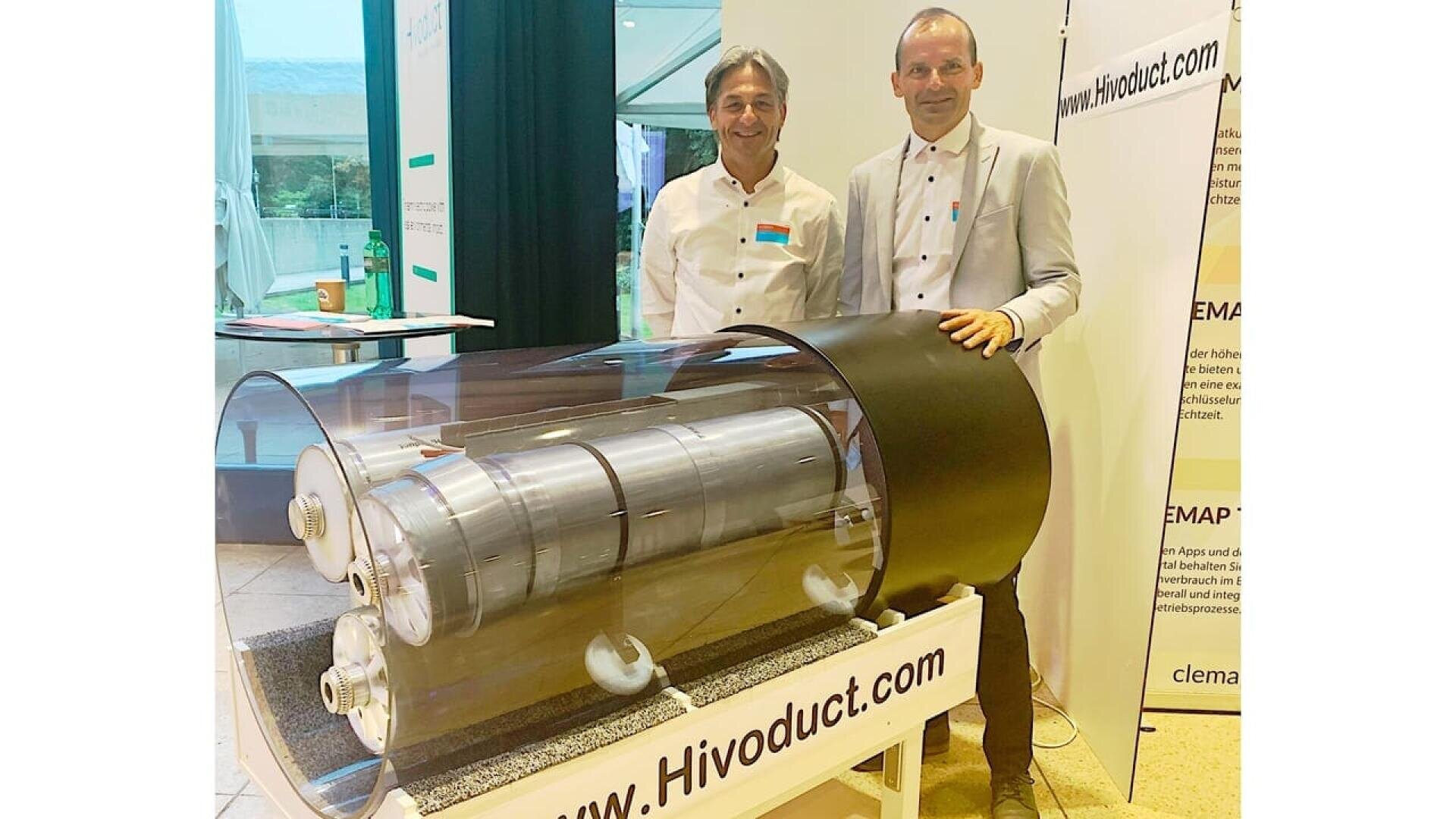 Two men in white shirts stand behind a cylindrical industrial device displayed on a stand labeled www.Hivoduct.com at an indoor exhibition or event with informational banners in the background.