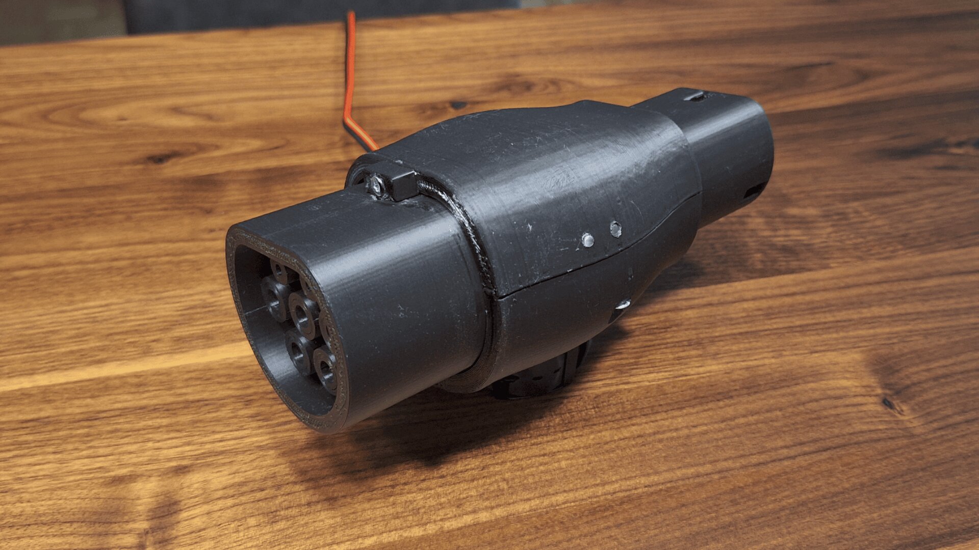 A black, cylindrical plastic device with a connector end and visible screws rests on a wooden table. A red wire is attached to the back of the device.