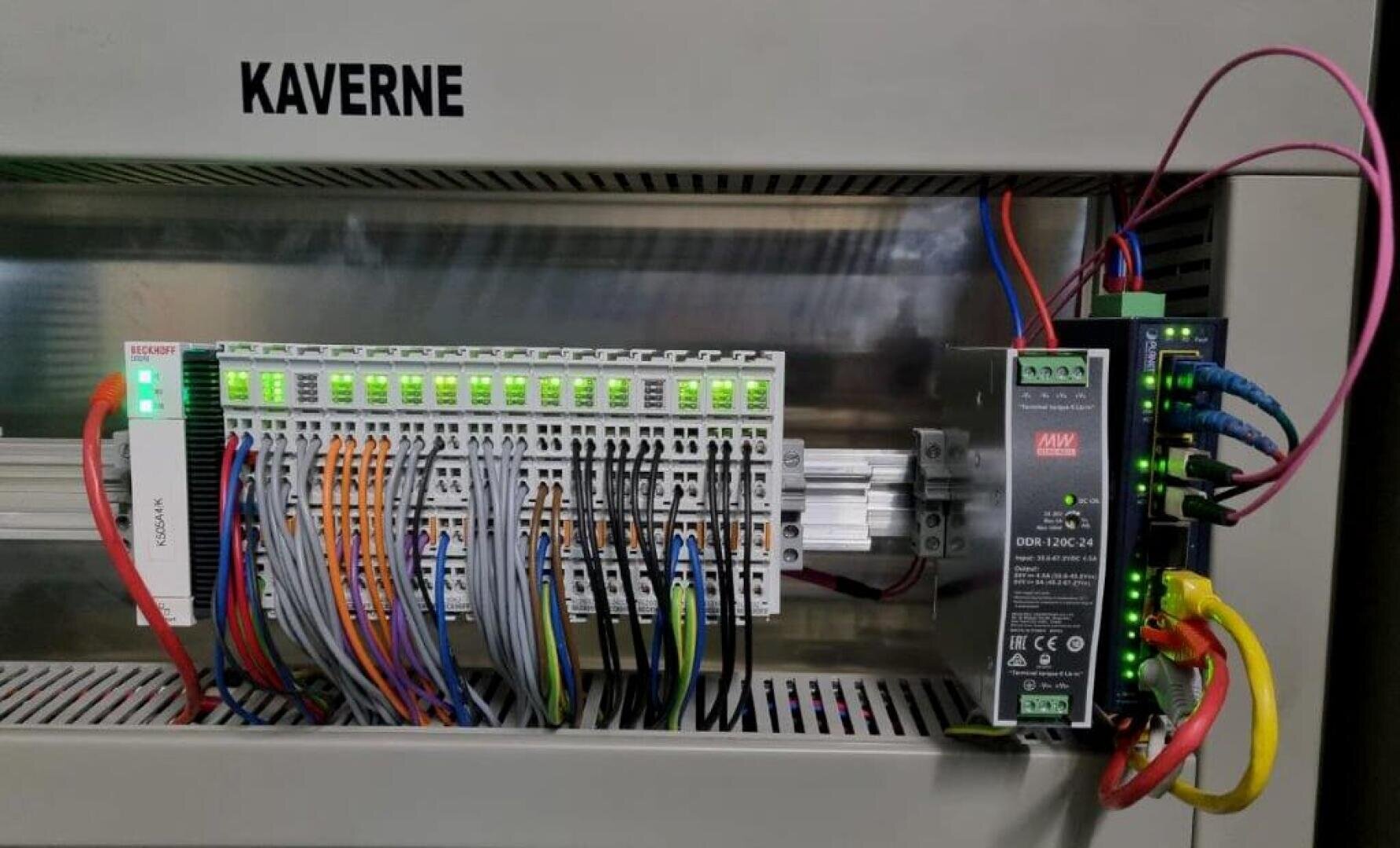 Ein mit KAVERNE beschriftetes Bedienfeld mit mehreren Signal- und Stromkabeln, die an Klemmenleisten und eine Stromversorgung angeschlossen sind, mit grünen Kontrollleuchten und verschiedenfarbigen Kabeln.