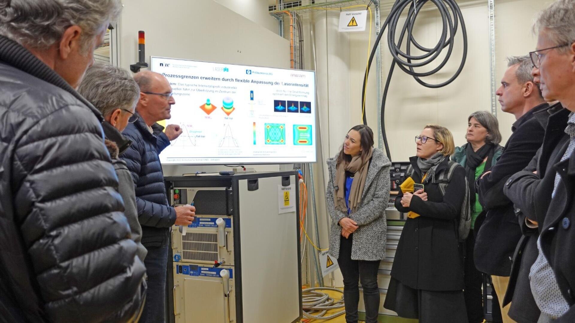 A group of people in a lab setting attentively listen to a man explaining information displayed on a large screen. Various cables and equipment are visible in the background.