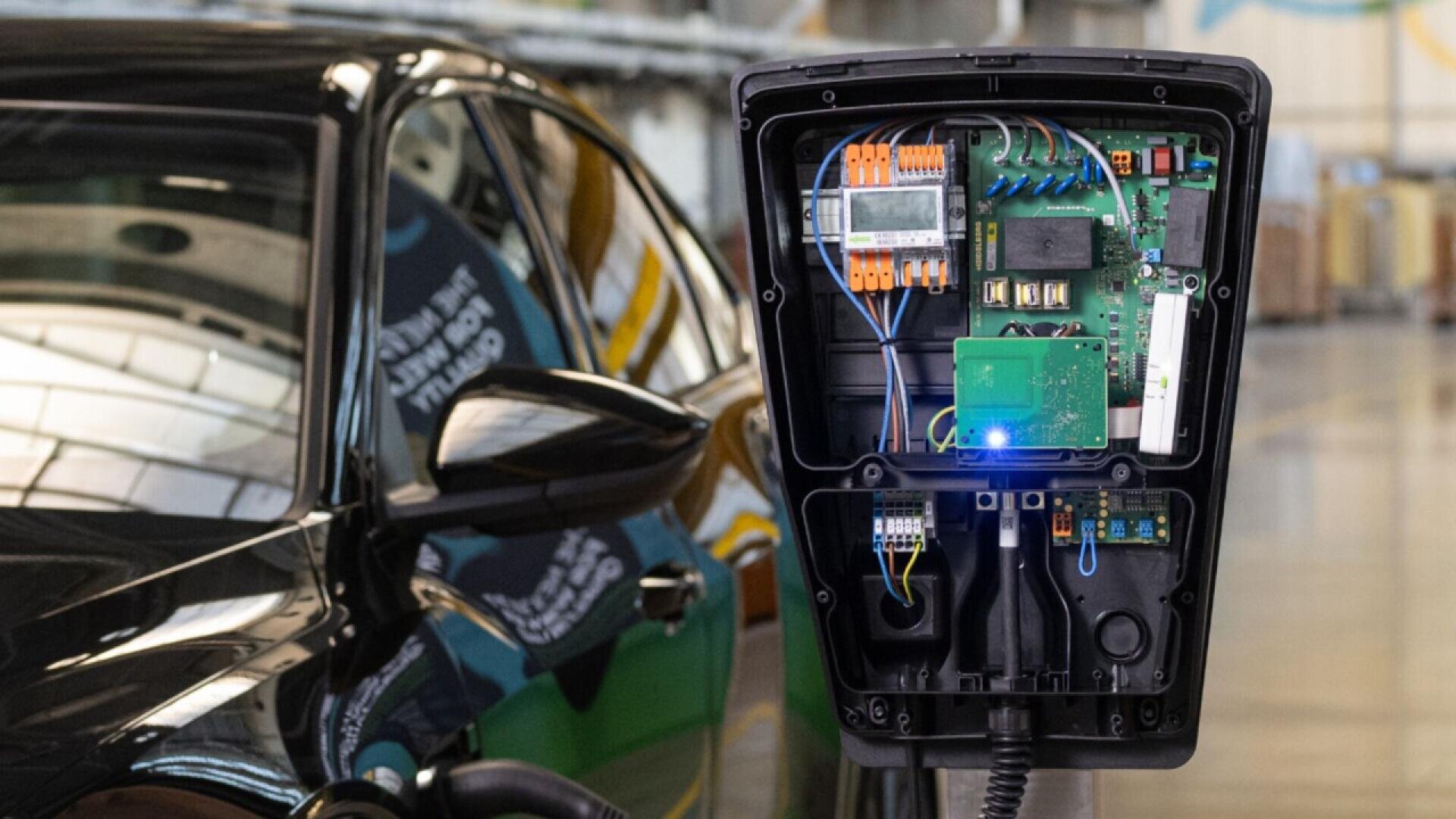 A close-up of an open electric vehicle charging station showing internal circuits and wiring, with a black car parked beside it inside an industrial building.