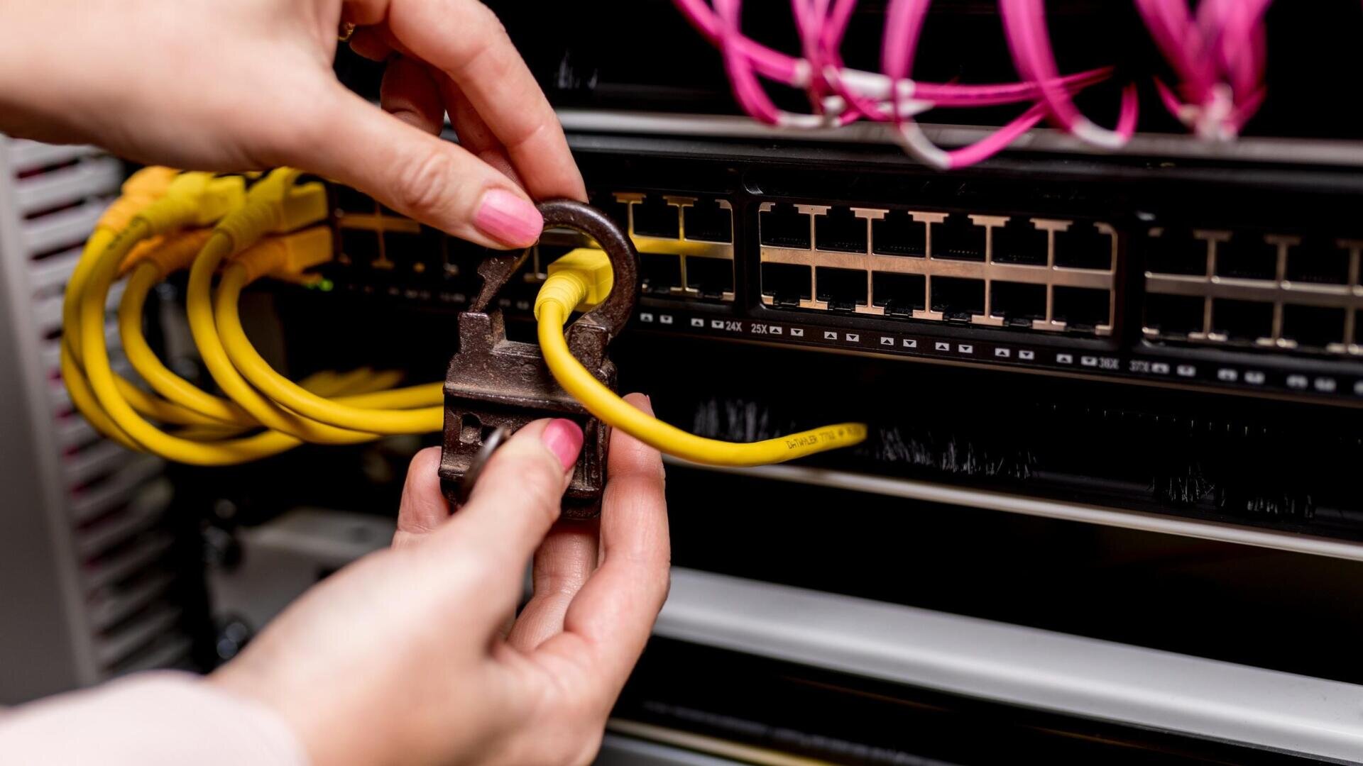 Die Hände einer Person, die ein gelbes Netzwerkkabel mit einem großen Vorhängeschloss an einem Serverschrank befestigt, als Symbol für Cybersicherheit oder Datenschutz. Im Hintergrund sind rosa und gelbe Kabel zu sehen.