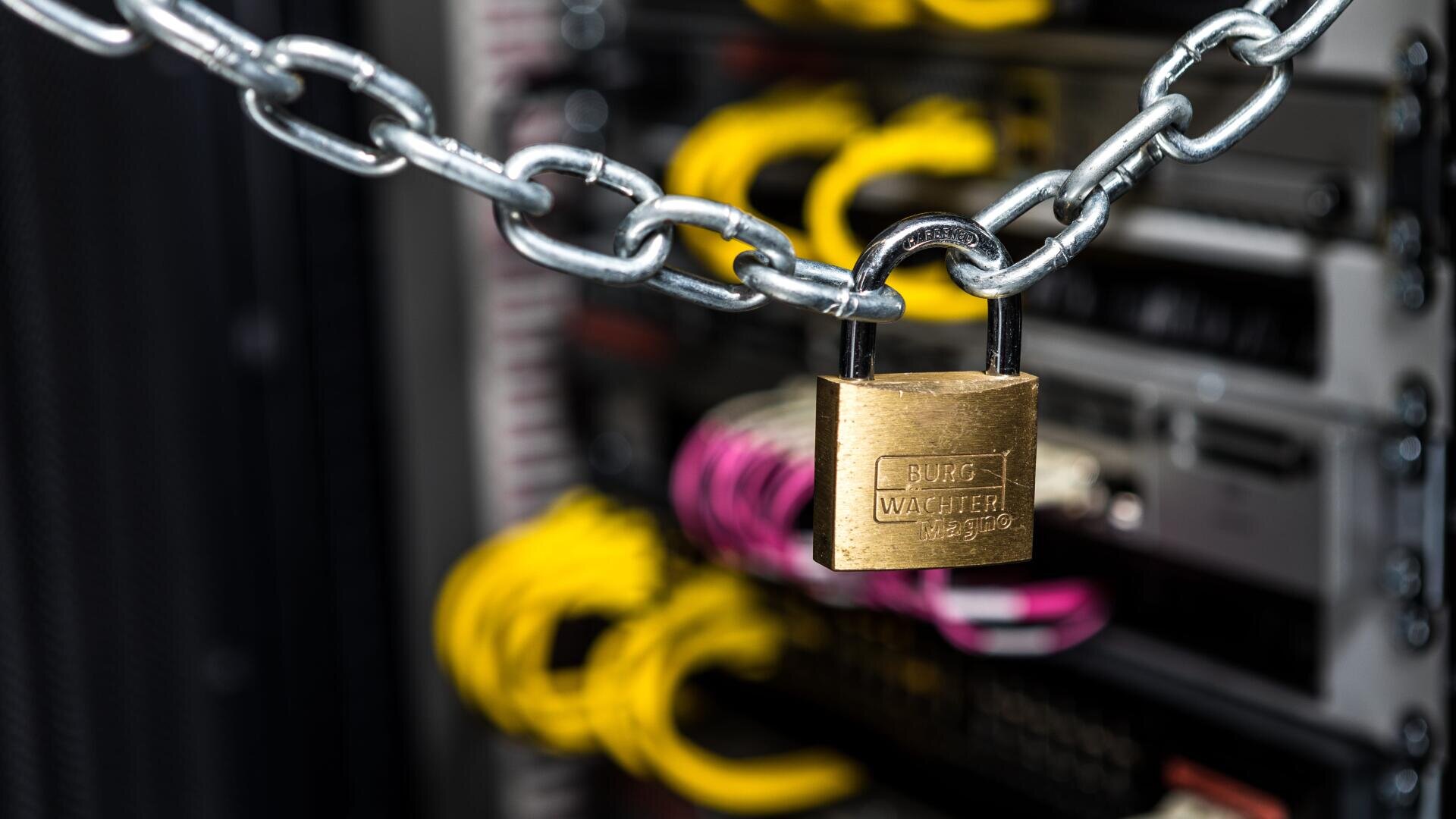 Eine Metallkette mit einem Vorhängeschloss aus Messing sichert ein Server-Rack und unterstreicht damit Cybersicherheit und Datenschutz. Im Hintergrund sind gelbe und rosa Netzwerkkabel zu sehen.