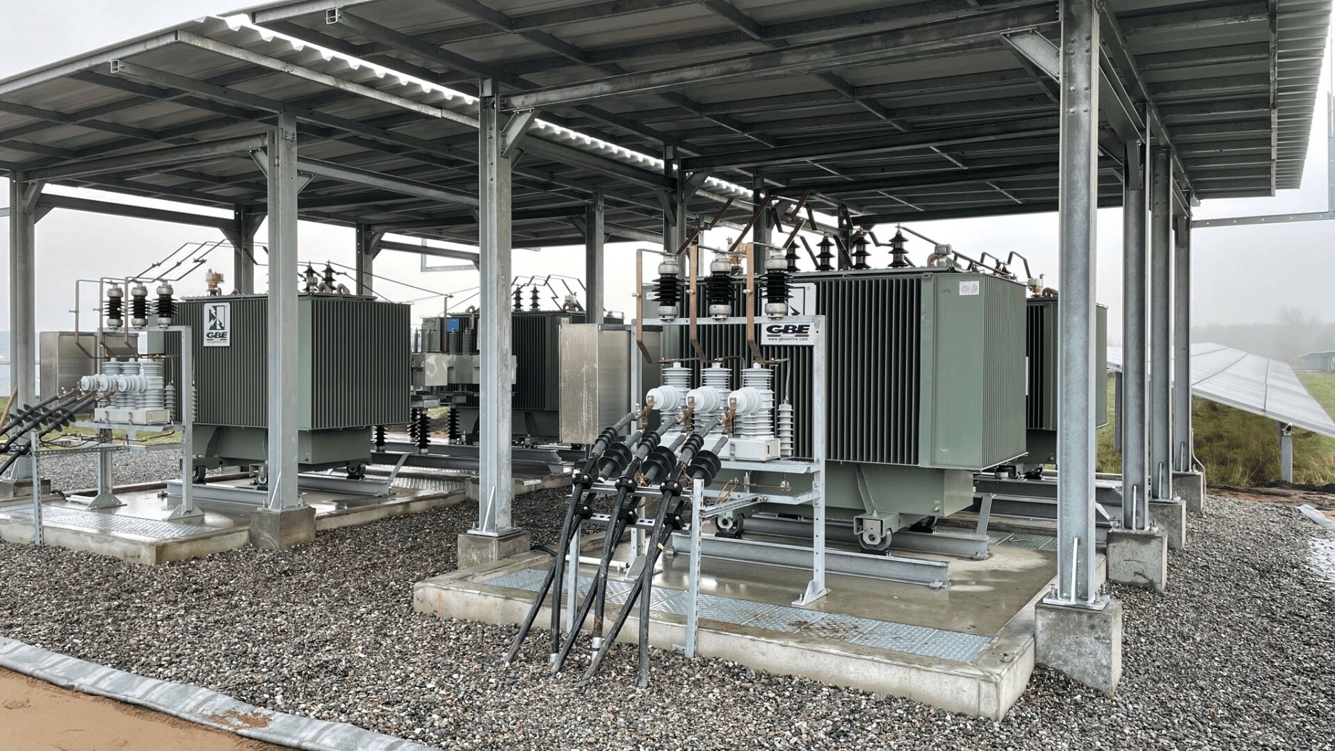 Two large electrical transformers are housed under a metal shelter on a gravel surface, with cables and electrical components connected. The area is outdoors with some grass and cloudy weather visible in the background.