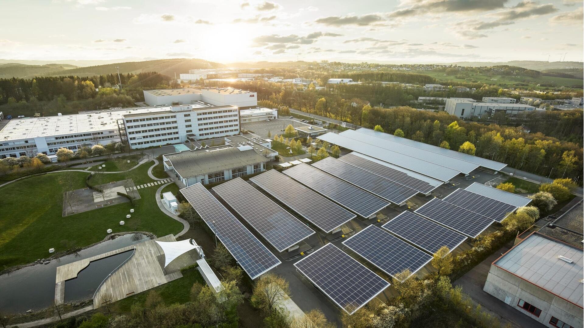 Luftaufnahme von großen Solarzellenfeldern auf Dächern und Parkhäusern eines Industrie- oder Bürokomplexes, umgeben von Bäumen und Gebäuden, mit der untergehenden Sonne im Hintergrund.