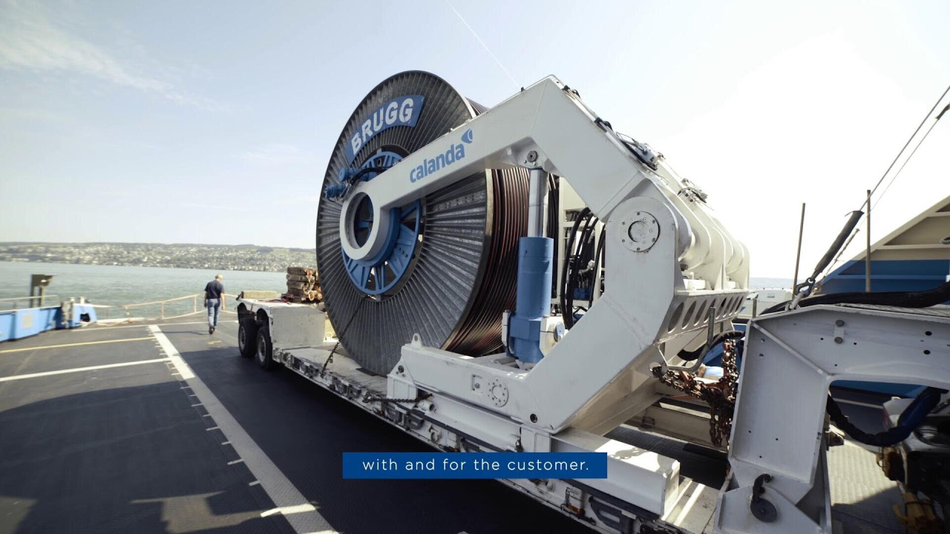 Eine große industrielle Kabeltrommel mit der Aufschrift BRUGG und callanda ist auf einer Maschine auf einem angedockten Schiff montiert, mit einem Gewässer und einer entfernten Küstenlinie im Hintergrund.
