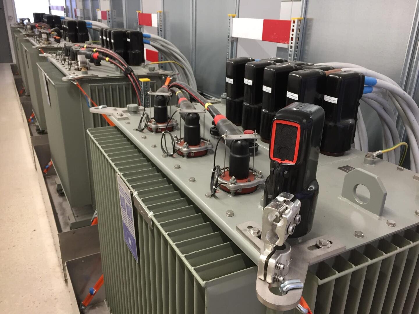 Eine Reihe elektrischer Transformatoren mit oben angebrachten Kabeln und Bauteilen, untergebracht in einer sauberen Halle mit grauen Metallwänden und -böden.