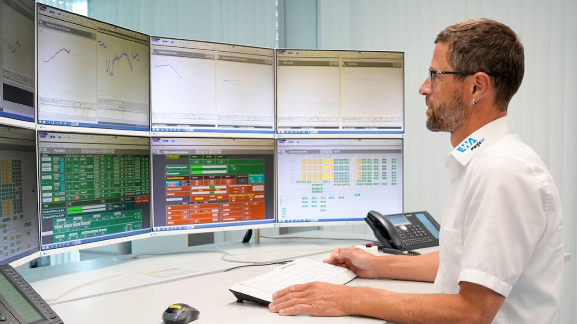 Ein Mann in weißer Uniform sitzt an einem Schreibtisch vor sechs großen Computermonitoren, die in einem modernen Kontrollraum Diagramme, Datentabellen und Grafiken anzeigen. Er tippt auf einer Tastatur und konzentriert sich auf die Bildschirme.