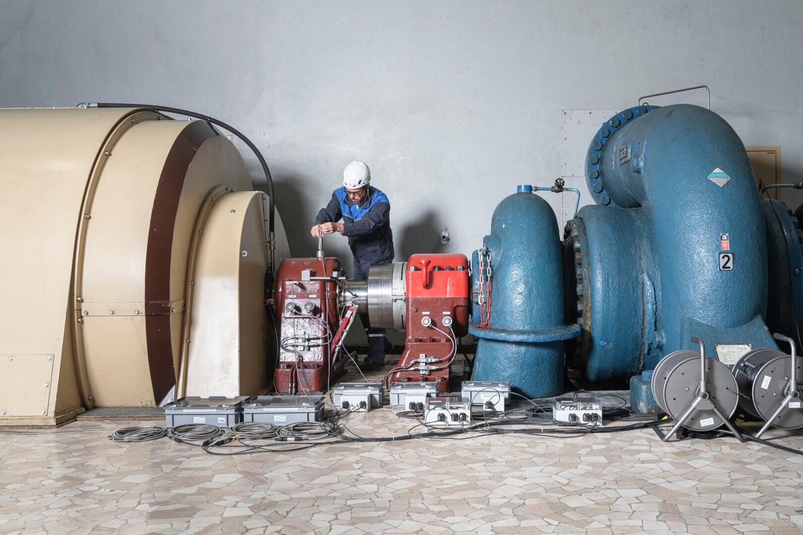 Ein Ingenieur mit weißem Helm arbeitet an Maschinen in einer Industrieanlage, umgeben von großen blauen und beigen mechanischen Geräten und Schaltkästen auf einem gefliesten Boden.