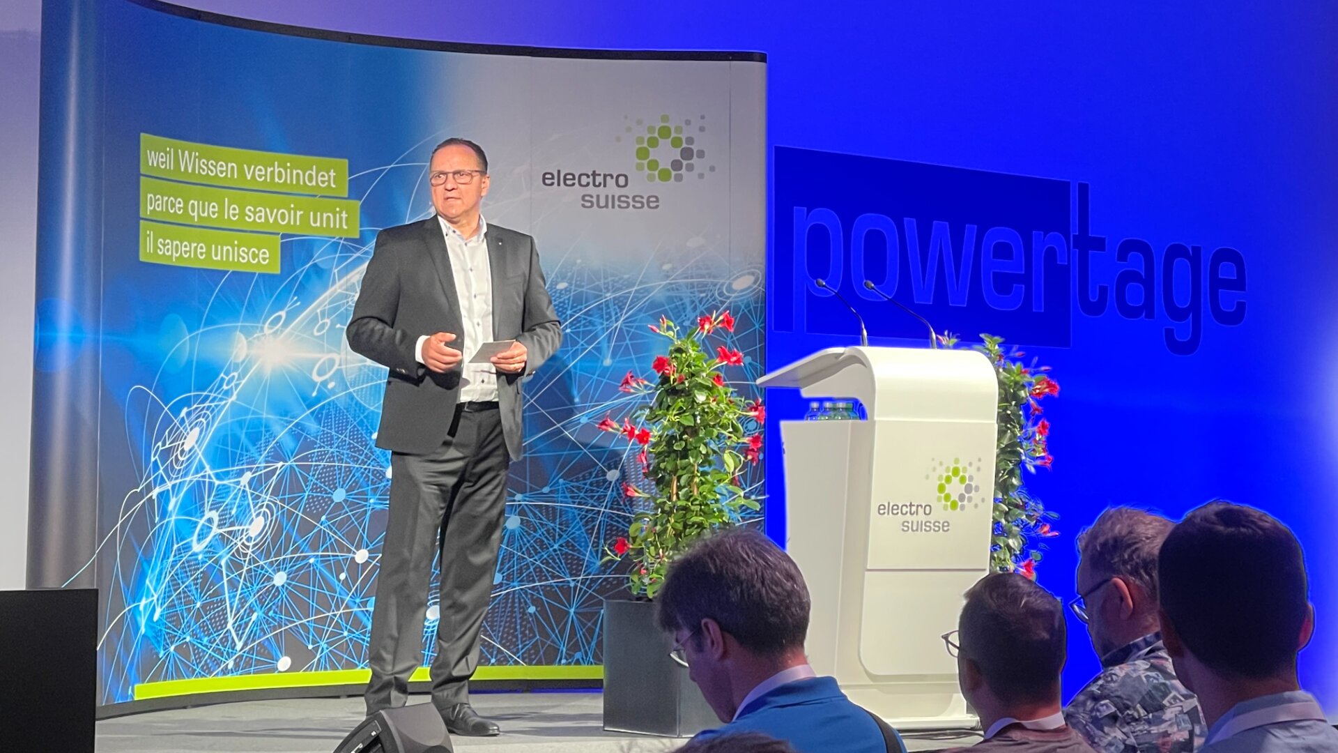 Ein Mann im Anzug spricht auf der Bühne einer Electro Suisse-Veranstaltung, vor einem Podium, Blumen und einem Hintergrund mit mehrsprachigem Text und Netzwerkgrafiken. Im Vordergrund sitzen die Zuhörerinnen und Zuhörer.