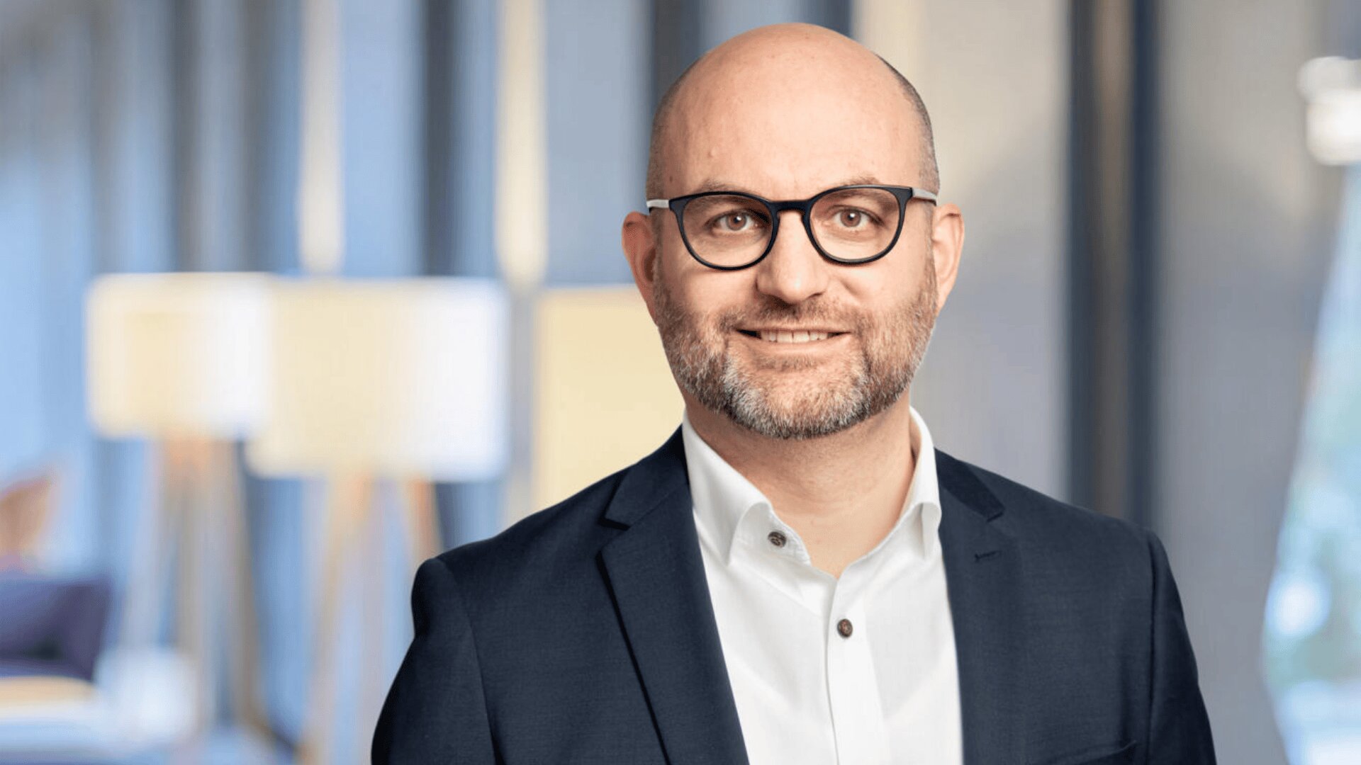 Ein lächelnder Mann mit Brille, kurzem Bart und Glatze, der ein weißes Hemd und einen dunklen Blazer trägt, steht in einem Innenraum mit weicher Beleuchtung und unscharfen Lampen im Hintergrund.