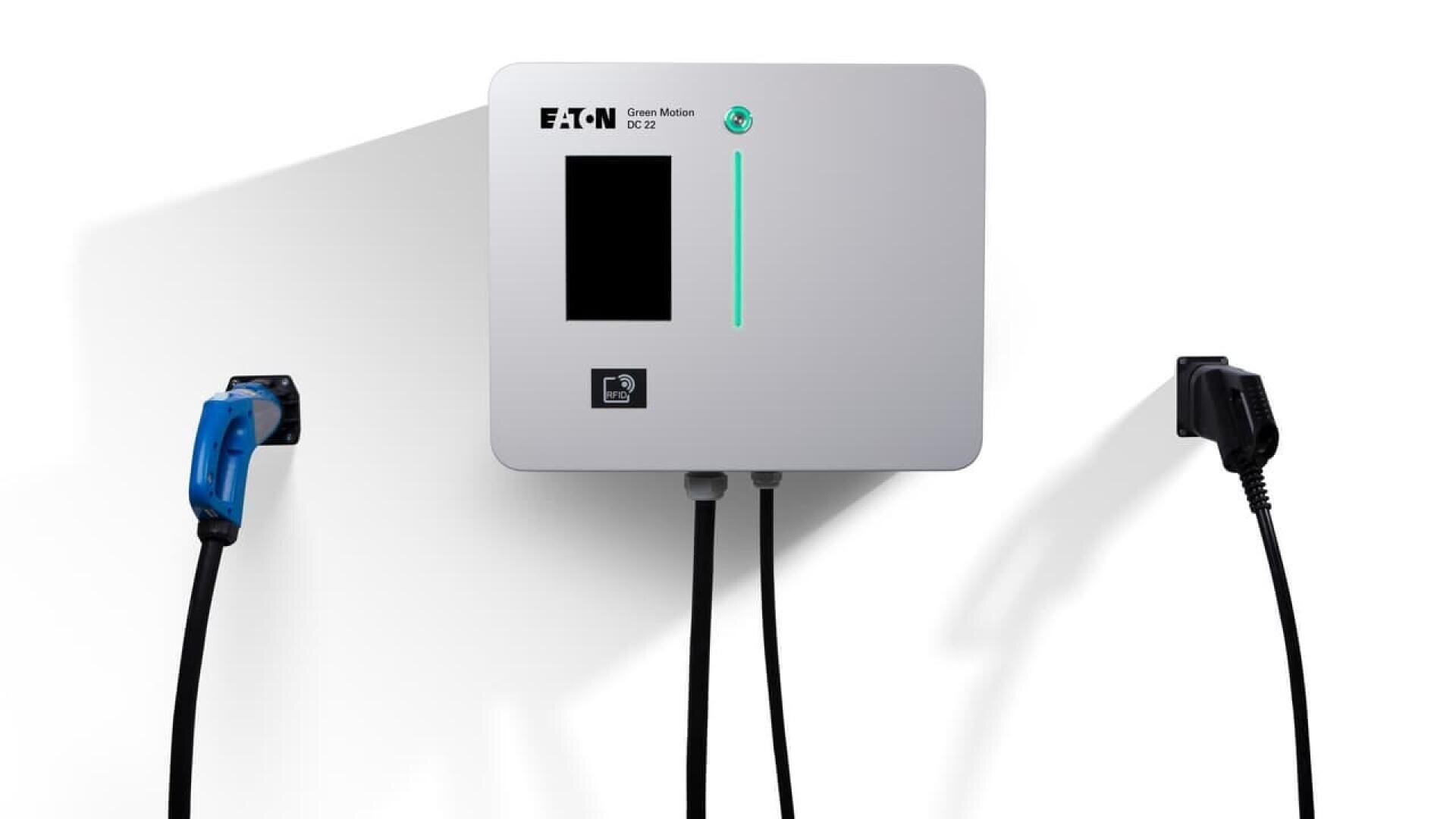 A wall-mounted Eaton Green Motion EV charger with a display screen, card reader, and two charging cables—one blue and one black—plugged into either side, casting shadows on a white background.