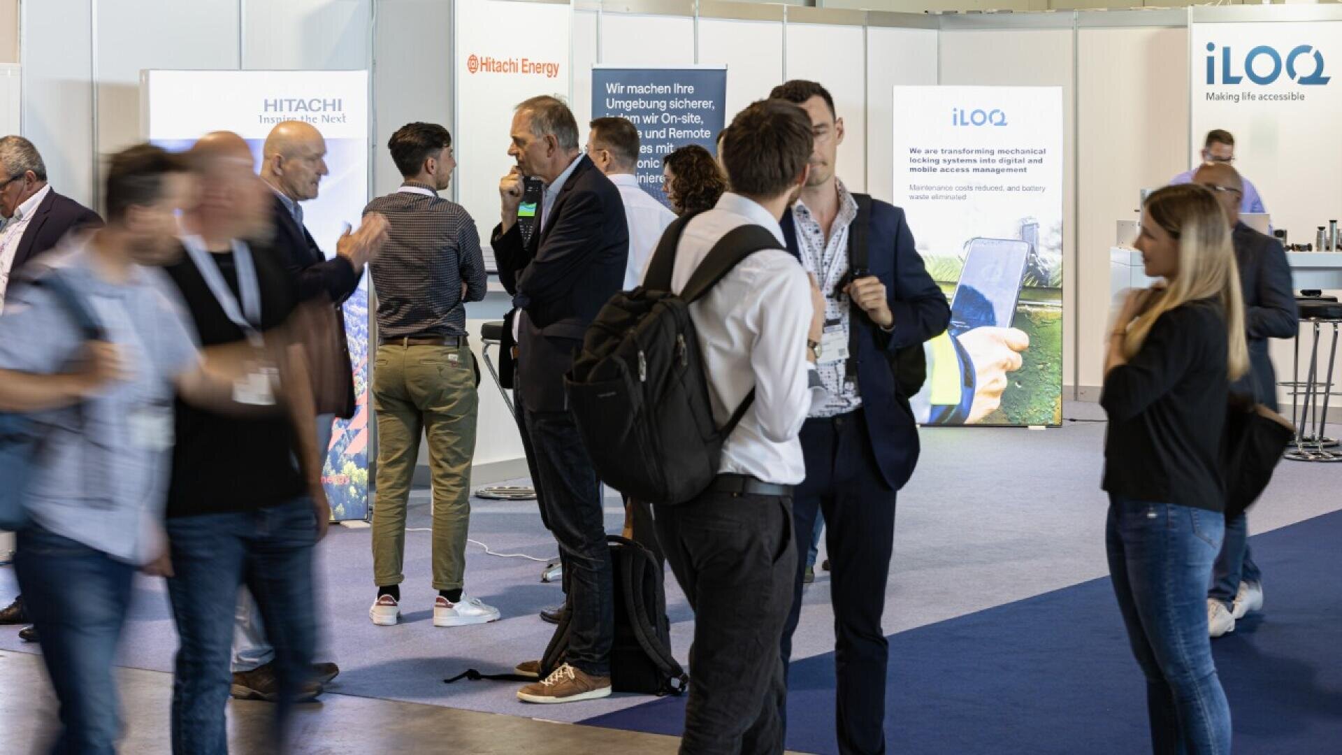 People are standing and talking in groups at a technology expo or conference, with company booths and banners visible in the background. Some people hold bags or wear lanyards with badges.