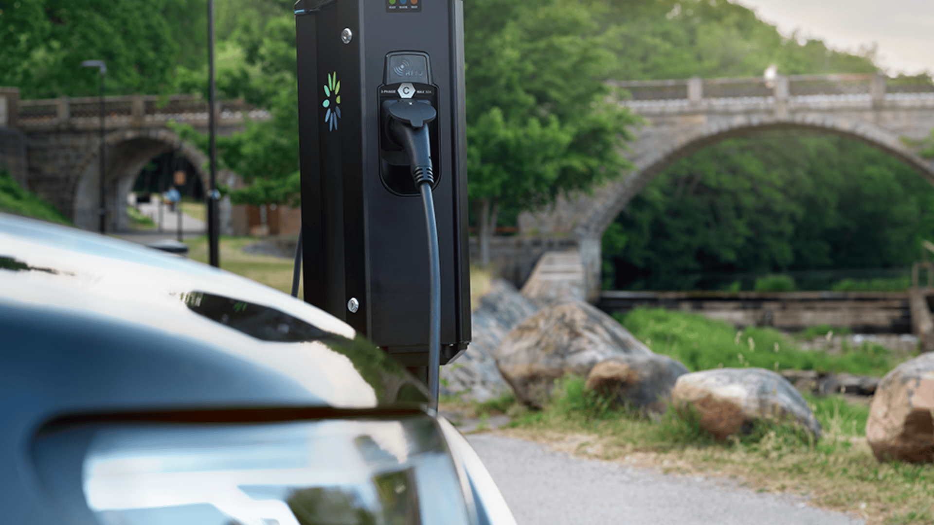 Ein Elektrofahrzeug wird an einer öffentlichen Ladestation in der Nähe einer Steinbrücke und grüner Bäume aufgeladen, mit großen Felsen und einer Grasfläche im Hintergrund.