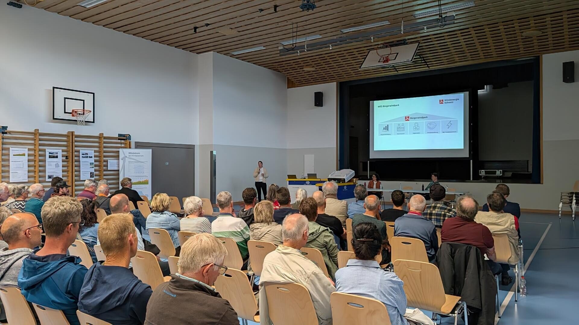 A group of people sitting in a sports hall in front of a stage where a presentation is being projected onto a screen. A speaker stands near the screen, and some participants are taking notes.