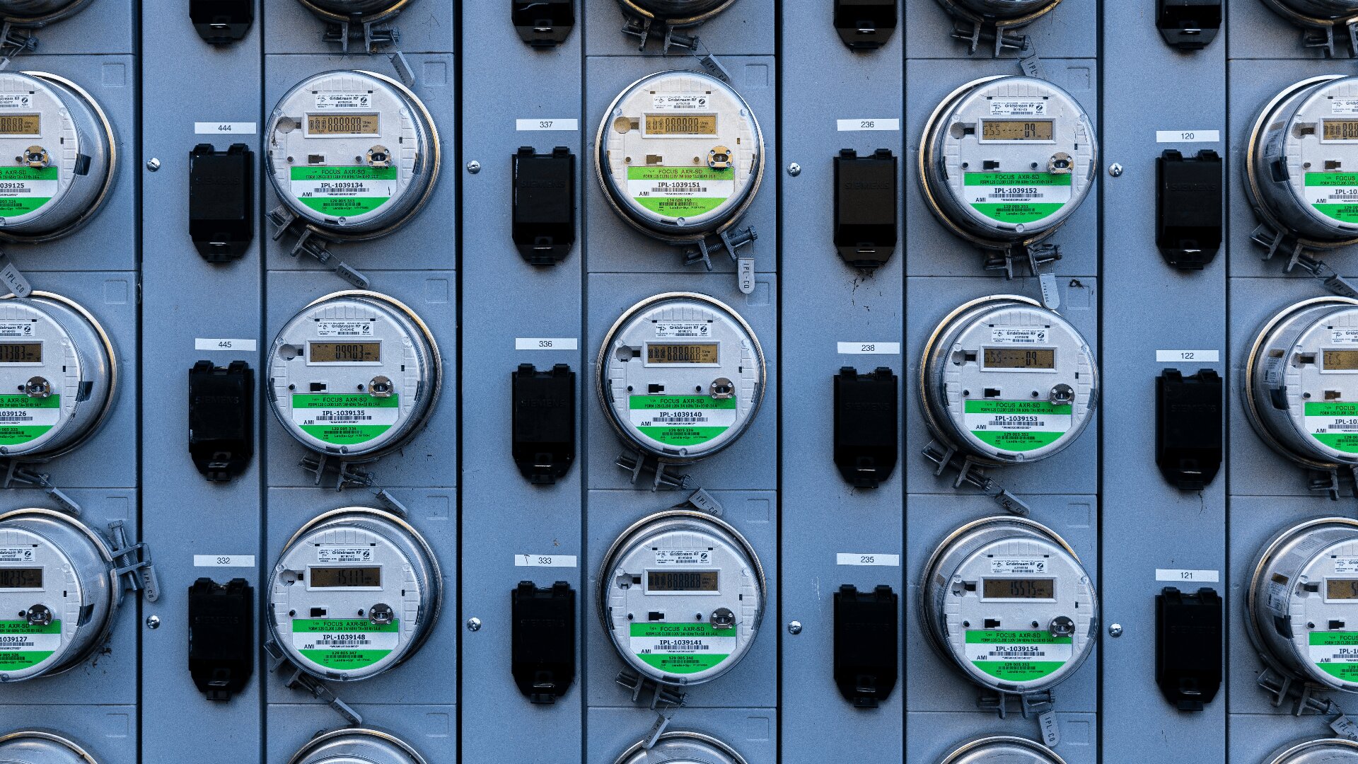 Ein Gitter aus mehreren Stromzählern, die an einer grauen Wand montiert sind, jeder mit digitaler Anzeige und grünen Etiketten, die einheitliche Reihen und Spalten zeigen.