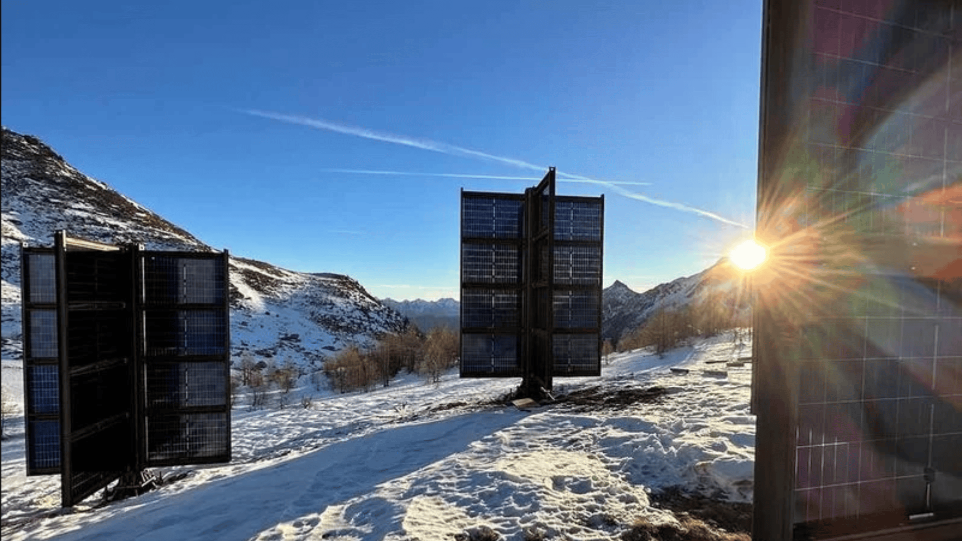 Solarmodule stehen in einer verschneiten Berglandschaft bei Sonnenaufgang oder Sonnenuntergang, wobei das Sonnenlicht durch eines der Module strömt und die Szene beleuchtet. Im Hintergrund sind schroffe Berge und ein klarer blauer Himmel zu sehen.