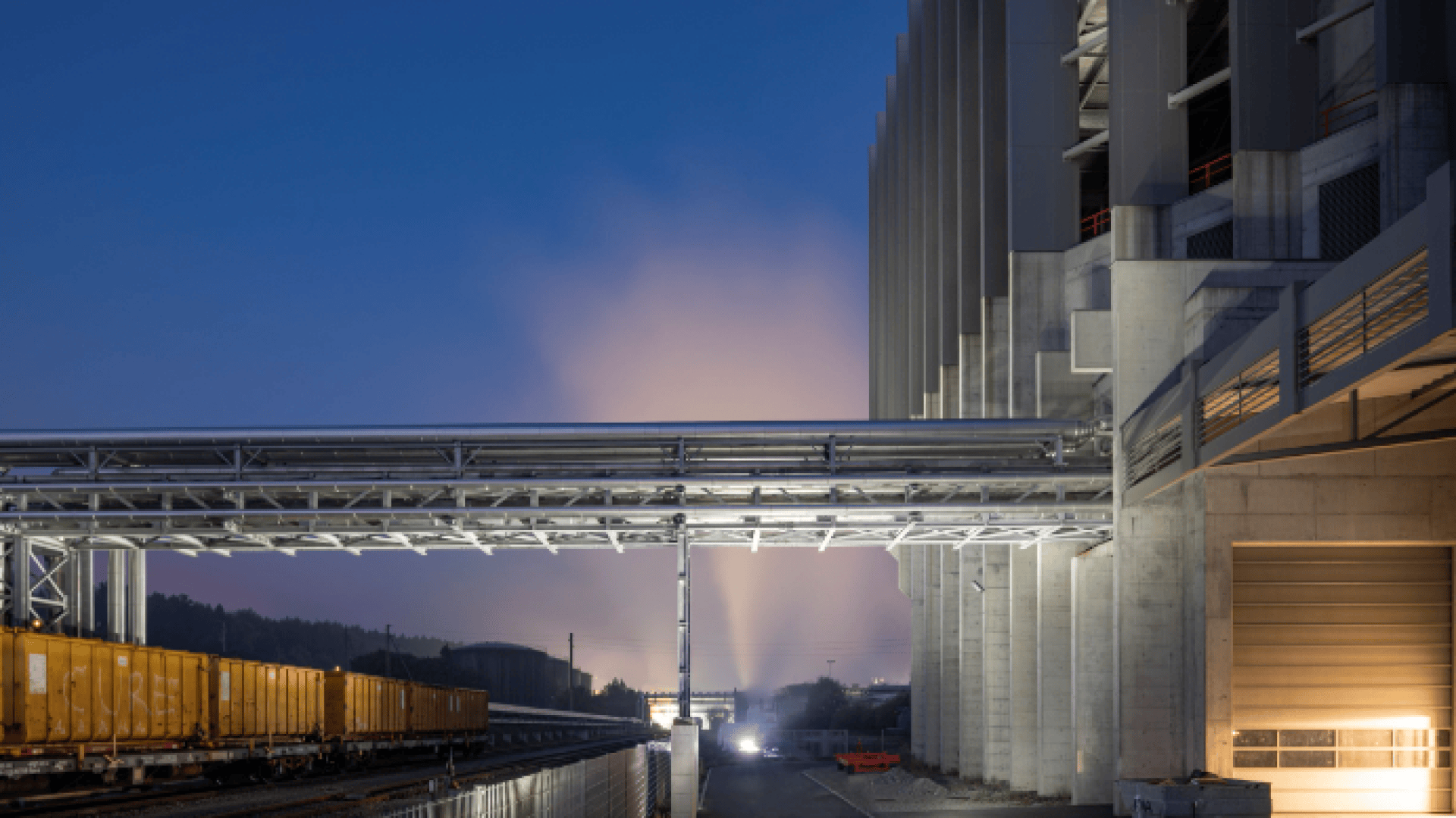 Industrieanlage in der Abenddämmerung mit beleuchtetem Rauch oder aufsteigendem Dampf im Hintergrund, Eisenbahnschienen und gelben Güterwagen auf der linken Seite und einem großen Betongebäude mit Metallstegen auf der rechten Seite.