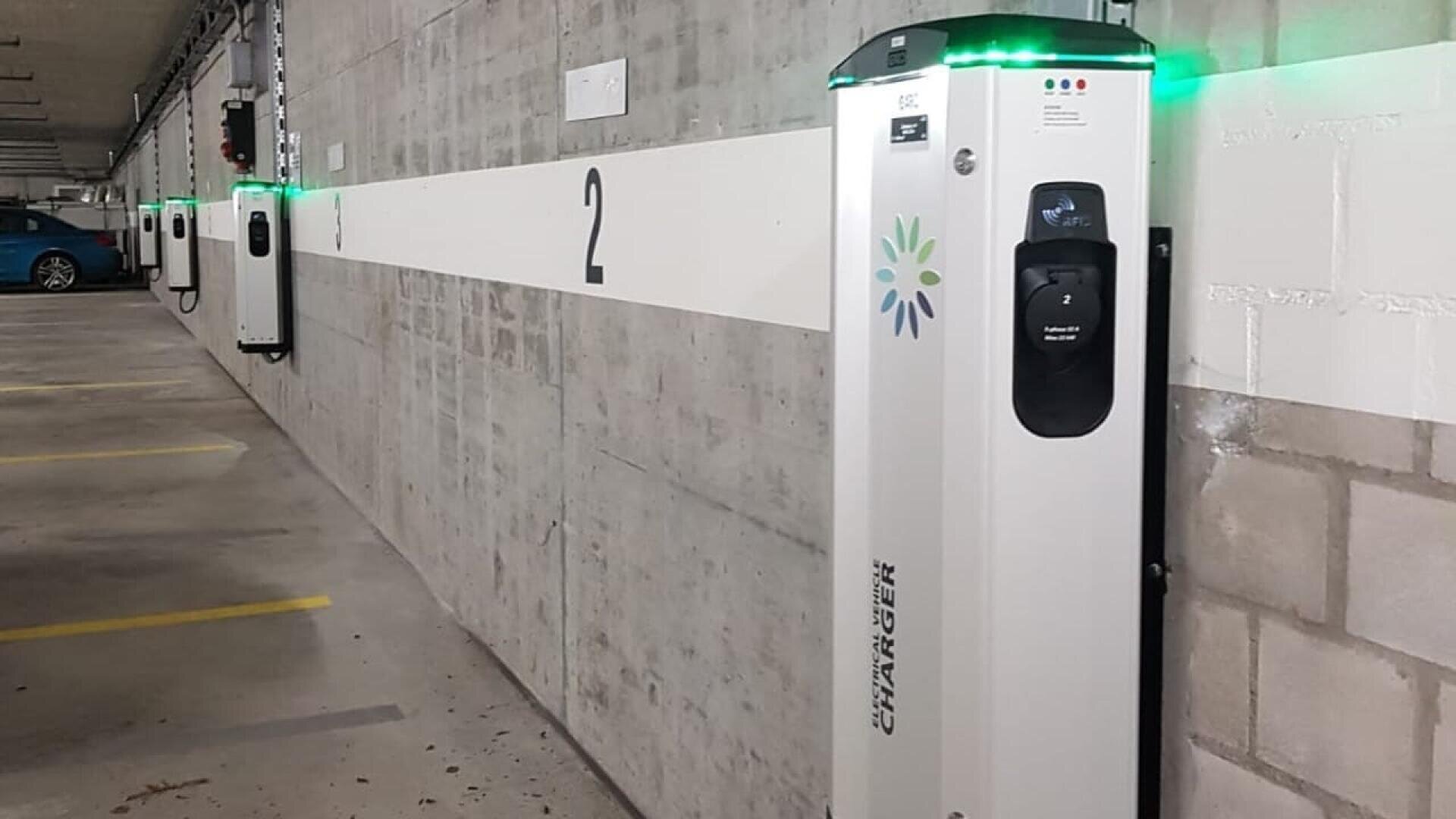 A row of electric vehicle charging stations is mounted on a concrete wall in an indoor parking garage, with green indicator lights on top. Yellow parking space lines are visible on the floor.