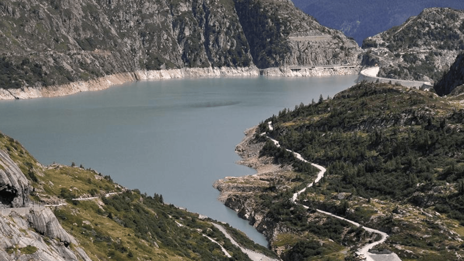 Eine kurvenreiche Straße führt an einem grünen, felsigen Berghang hinunter zu einem großen See, der von steilen Klippen und bewaldeten Hängen umgeben ist, wobei in der Ferne ein Damm zu sehen ist.