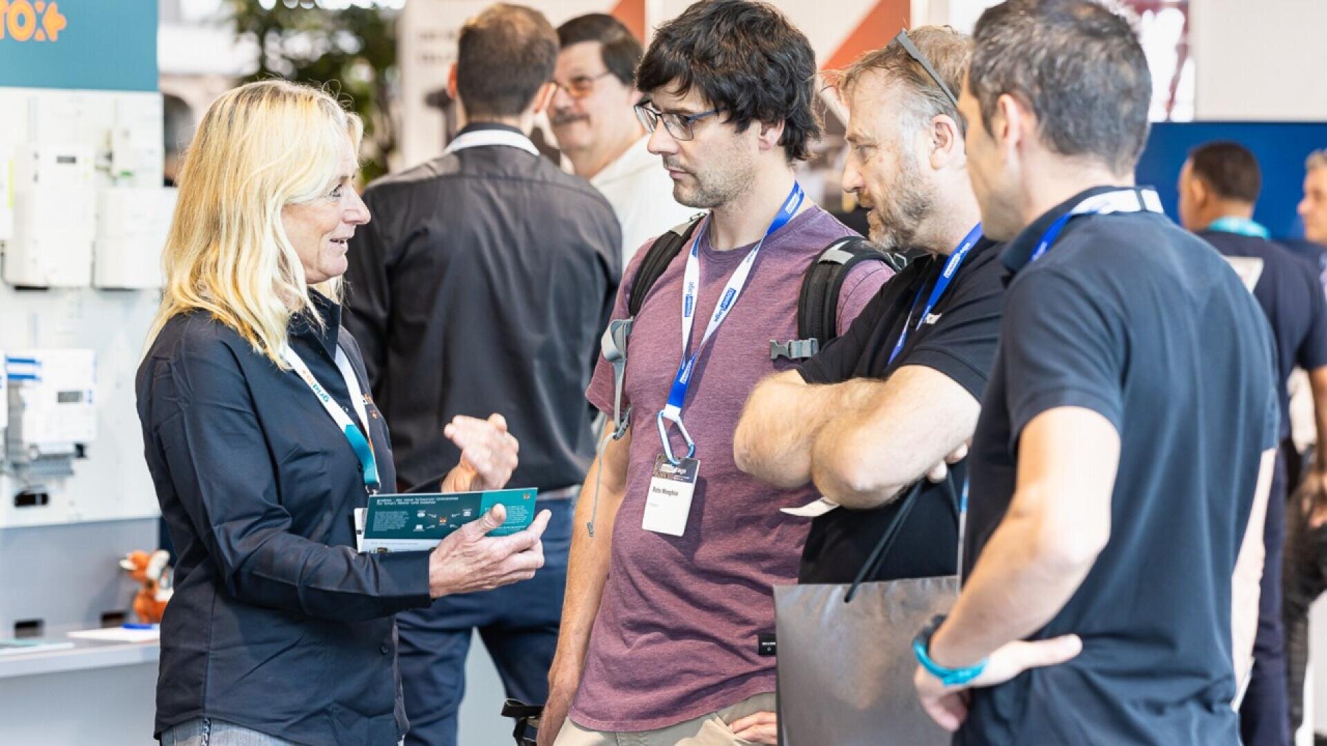 A woman holding a brochure speaks to three men wearing lanyards at an indoor event or conference. The group appears engaged in conversation, with other people and displays visible in the background.