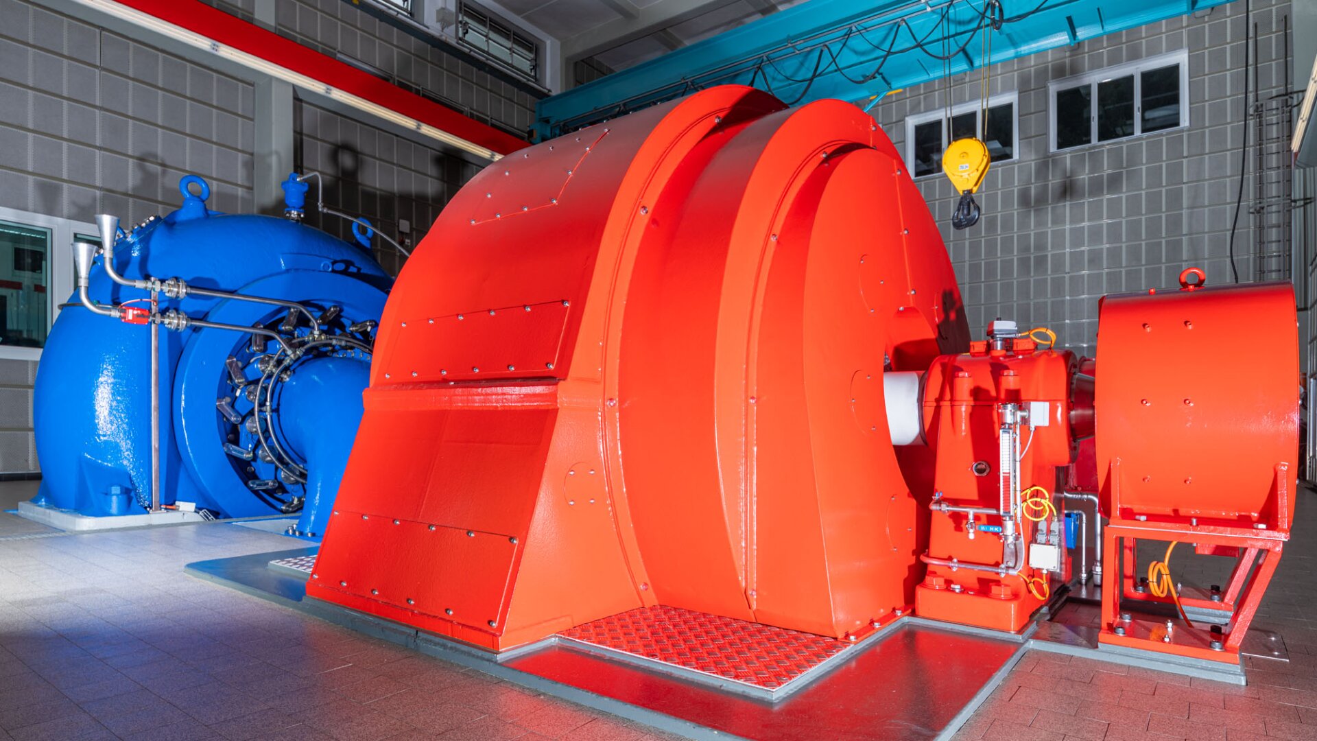Eine große Industriemaschine mit einem leuchtend roten Turbinengehäuse und einer blauen Generatoreinheit ist in einem Innenraum mit grau gekachelten Wänden und Deckenbeleuchtung installiert.