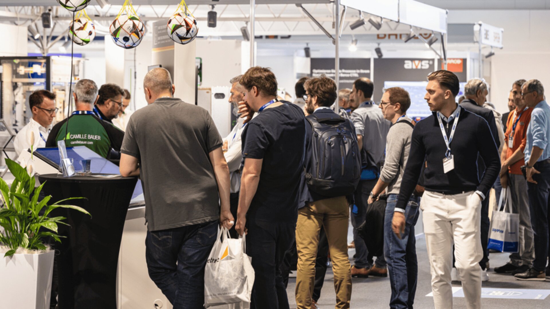Eine Gruppe von Menschen steht in einer Reihe und geht auf einer belebten Messe oder Ausstellung umher, wobei im Hintergrund verschiedene Ausstellungsstände, Banner und hängende Fußbälle zu sehen sind.