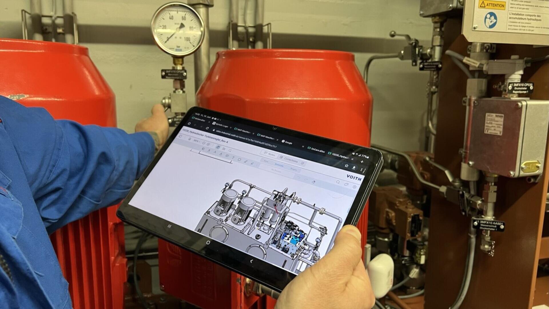 A person holds a tablet displaying a technical diagram in front of industrial equipment, including red tanks and a pressure gauge, in what appears to be a mechanical or engineering setting.