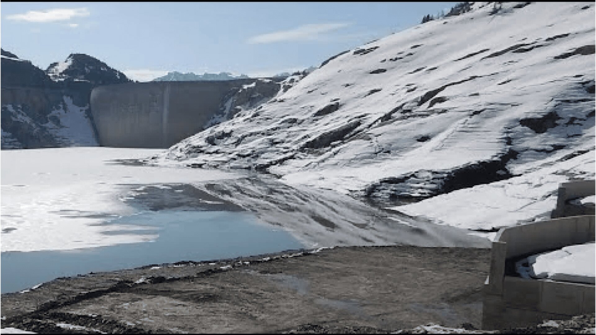 Ein großer Staudamm steht am Ende eines teilweise zugefrorenen Sees, der von schneebedeckten Bergen umgeben ist, mit einigen freiliegenden Felsen im Vordergrund.