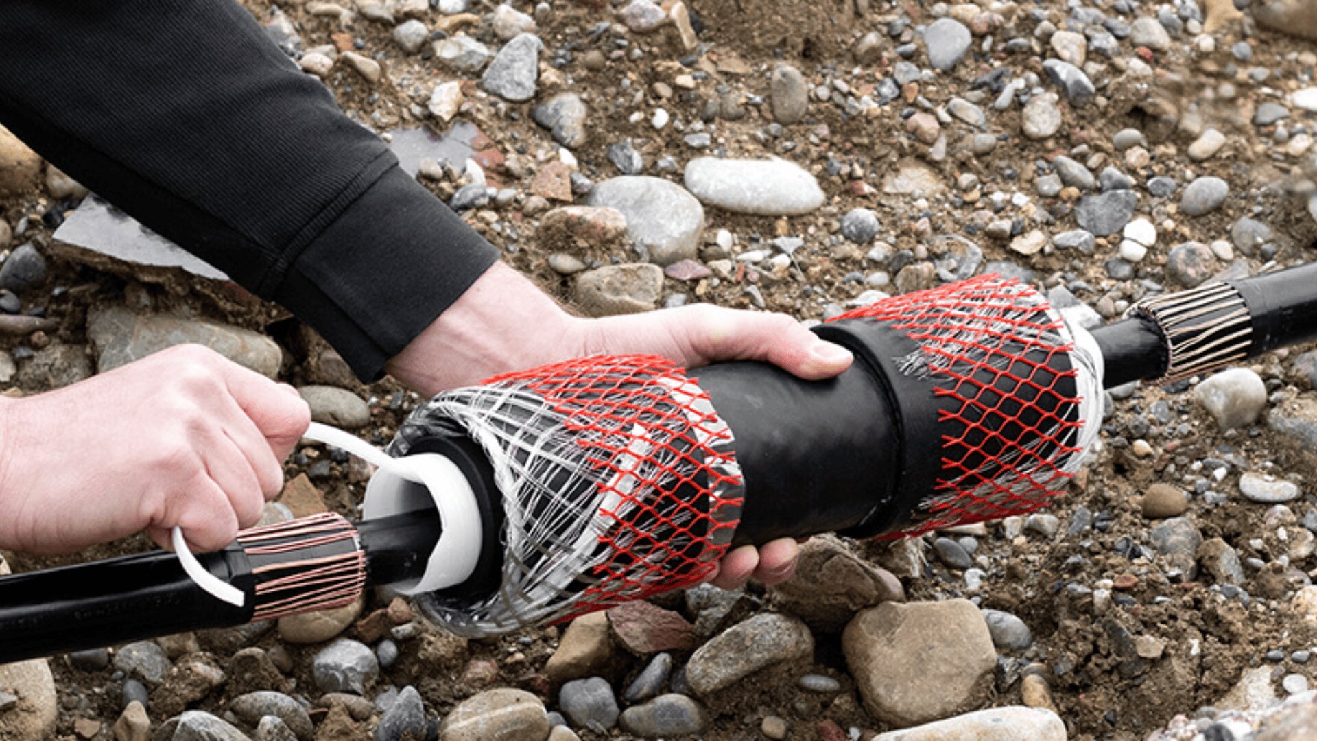 A person’s hands installing a protective splice cover over an underground cable, using red mesh sleeves for support, surrounded by dirt and rocks.