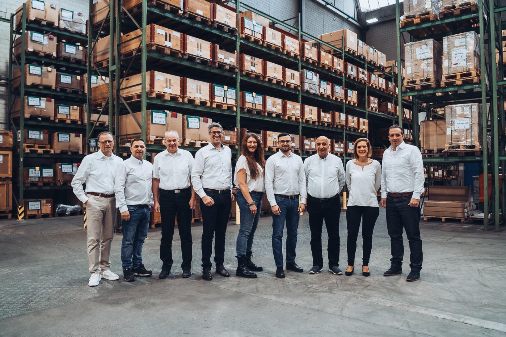 A group of ten people in business casual attire stand side by side, smiling at the camera. They are in a warehouse with high shelves stacked with boxes and pallets.