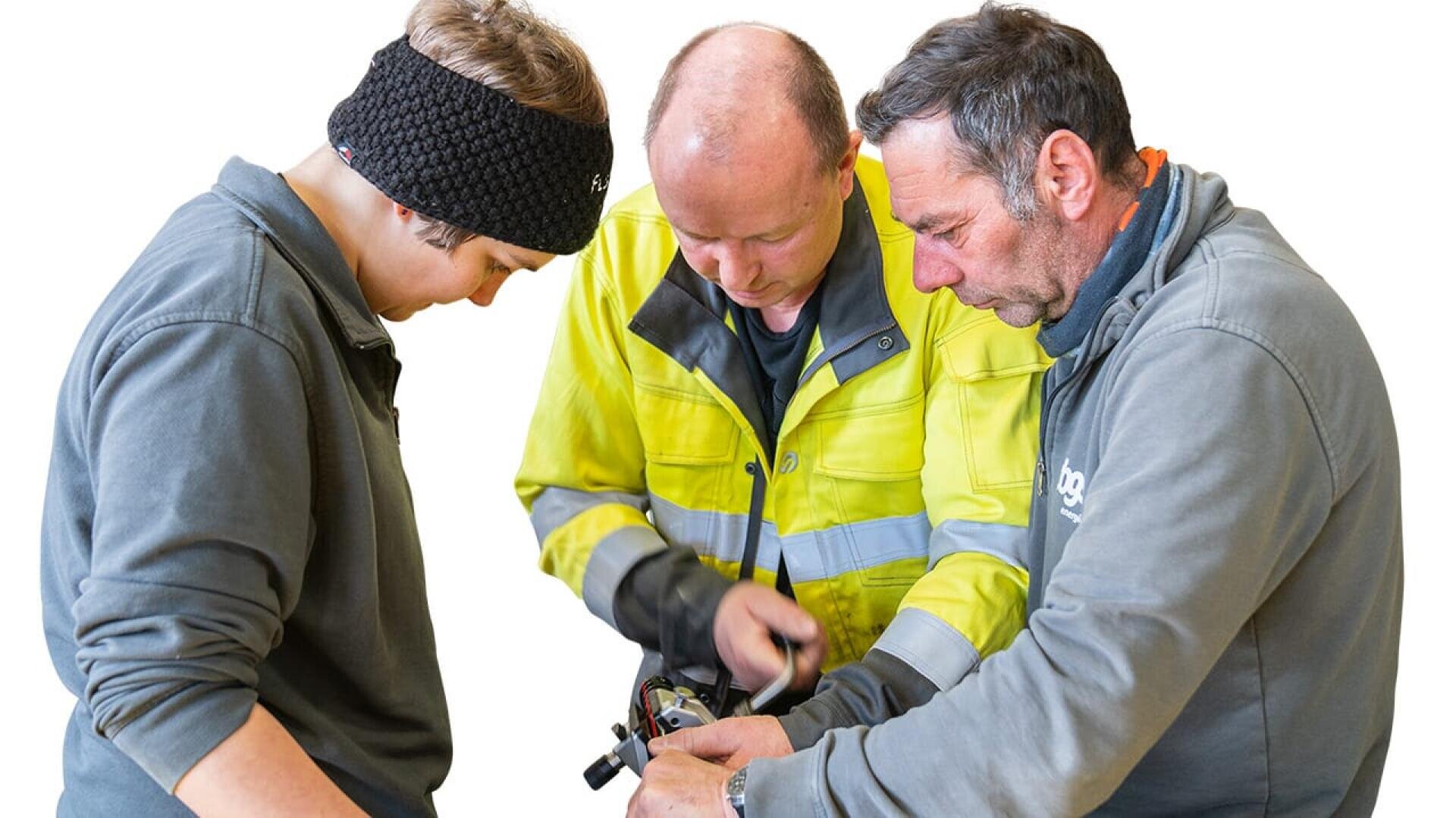Drei Personen in Arbeitsjacken untersuchen und bearbeiten gemeinsam ein mechanisches Gerät und konzentrieren sich dabei auf ihre Aufgabe. Der Hintergrund ist einfarbig weiß.