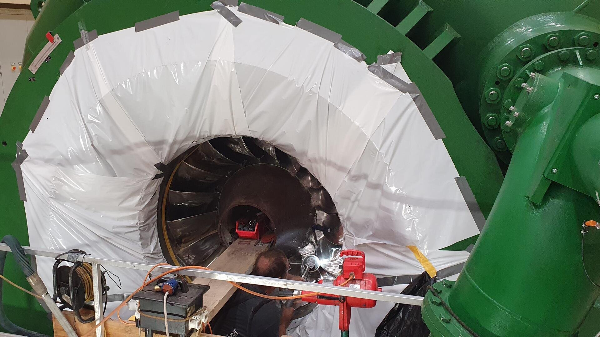 Eine Person arbeitet an dem großen freiliegenden Rotor einer grünen Industriemaschine, der teilweise mit weißer Plastikfolie abgedeckt und mit Klebeband gesichert ist. Werkzeuge und Ausrüstung liegen in der Nähe verstreut.