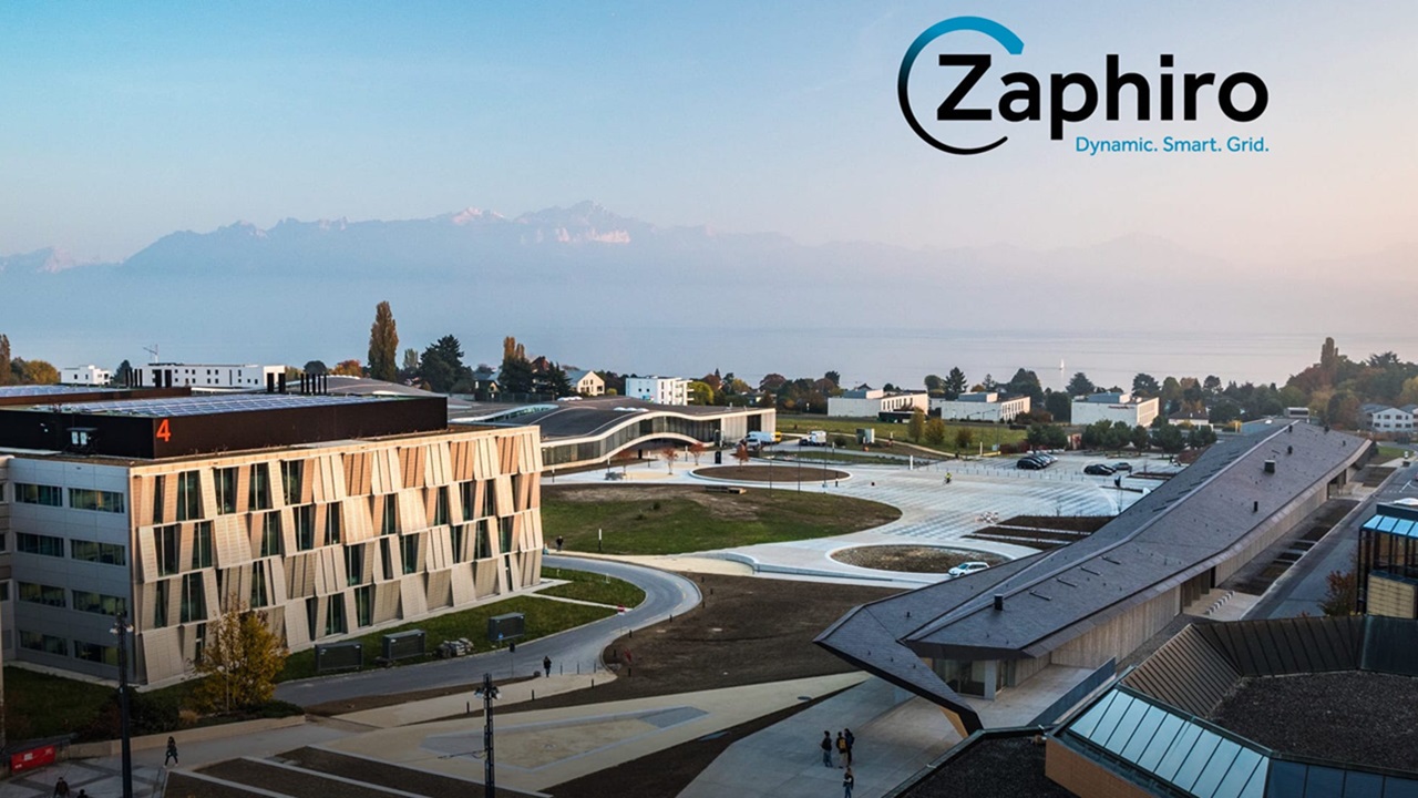 Ein moderner Campus mit zeitgenössischen Gebäuden, Grünflächen und einem Parkplatz. Das Zaphirio-Logo und der Slogan Dynamic. Smart. Grid. erscheinen im Himmel über der Szene, mit Bergen und einem See im Hintergrund.