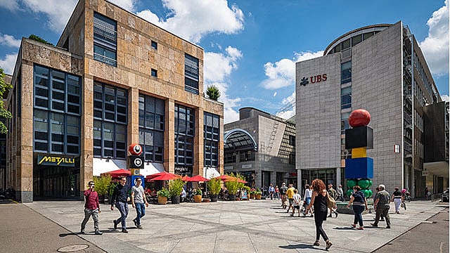 Ein lebhafter öffentlicher Platz mit Menschen, die im Freien spazieren gehen und essen, umgeben von modernen Gebäuden, darunter eine UBS-Bank und eine bunte, gestapelte Skulptur. Der Himmel ist blau mit Wolken.