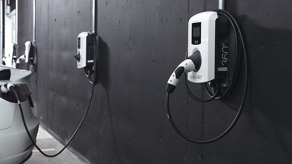 Ein Elektrofahrzeug ist an eine an der Wand befestigte Ladestation auf einem Parkplatz angeschlossen, eine weitere Ladestation ist weiter hinten an der schwarzen Wand zu sehen.