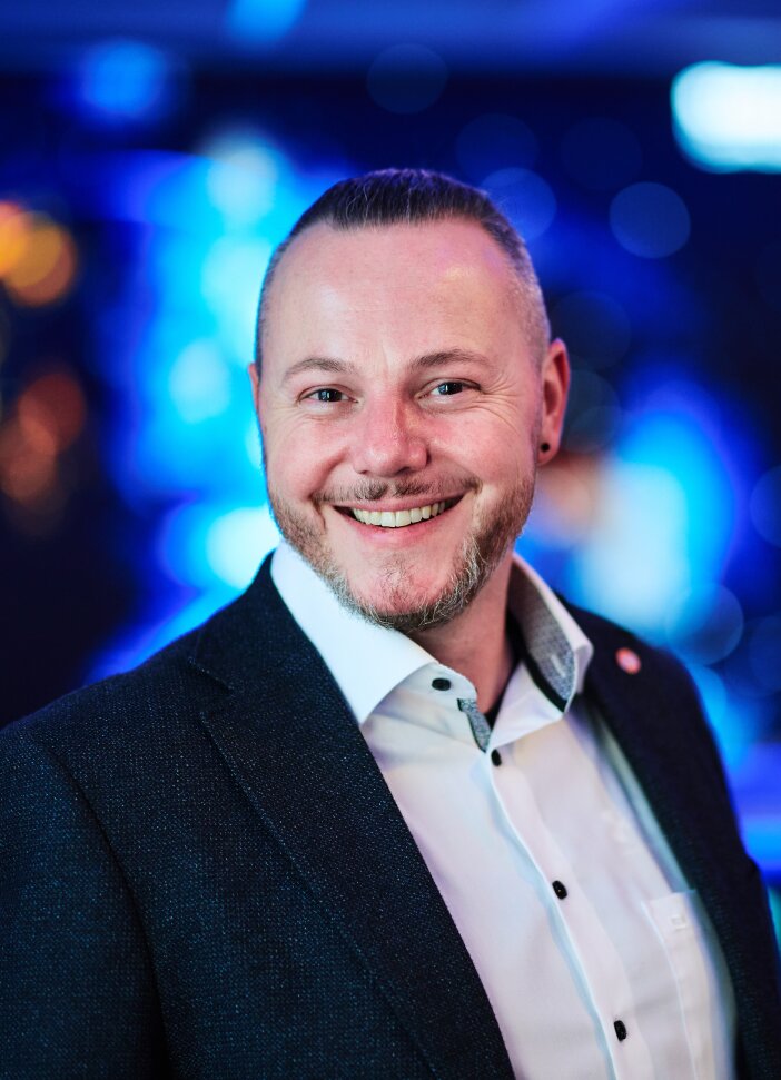 A smiling man with short hair and a trimmed beard, wearing a dark blazer over a white shirt, poses in front of a bright blue and purple blurred background.
