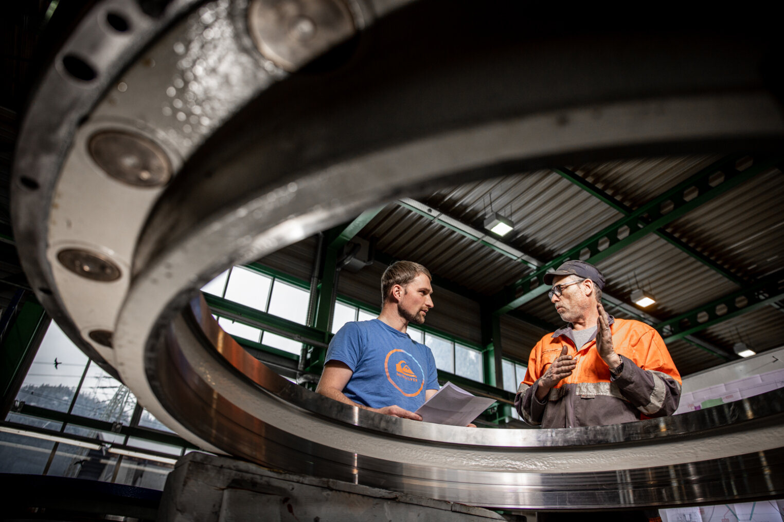 Zwei Männer stehen in einer großen Industrieanlage, die durch eine kreisförmige Metallstruktur zu sehen ist. Der eine trägt ein blaues Hemd und hält Papiere in der Hand, der andere trägt eine Sicherheitsjacke und eine Mütze.