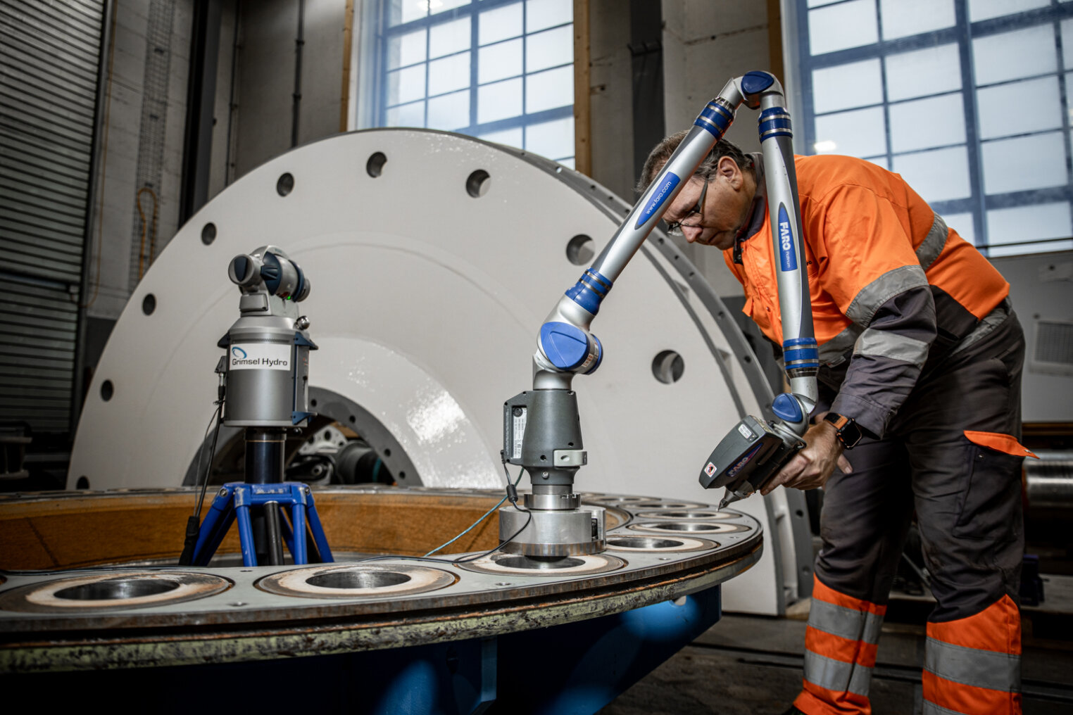 Ein Mann in einer orangefarbenen Sicherheitsjacke benutzt einen tragbaren 3D-Messarm, um ein großes industrielles Metallteil in einer Fabrikumgebung mit Maschinen und großen Fenstern im Hintergrund zu scannen.