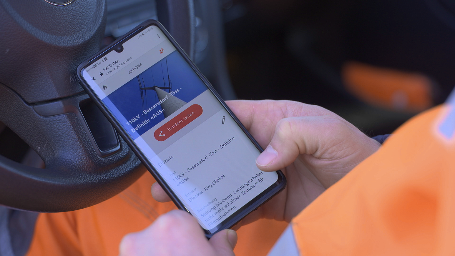 A person in an orange jacket uses a smartphone while sitting in a car. The phone screen displays an app with information about an electric vehicle charging station.